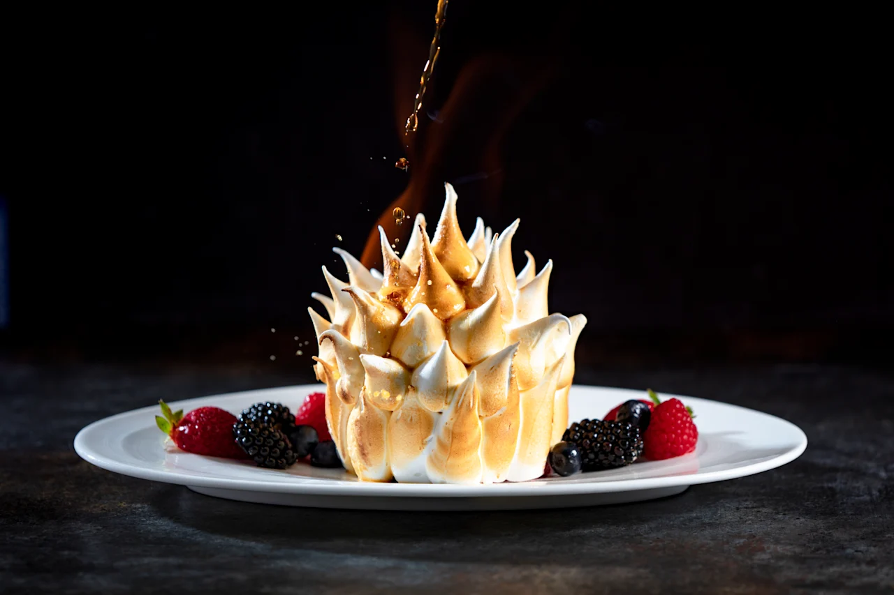 Toasted meringue dessert with fresh berries on white plate, caramel being drizzled on top.