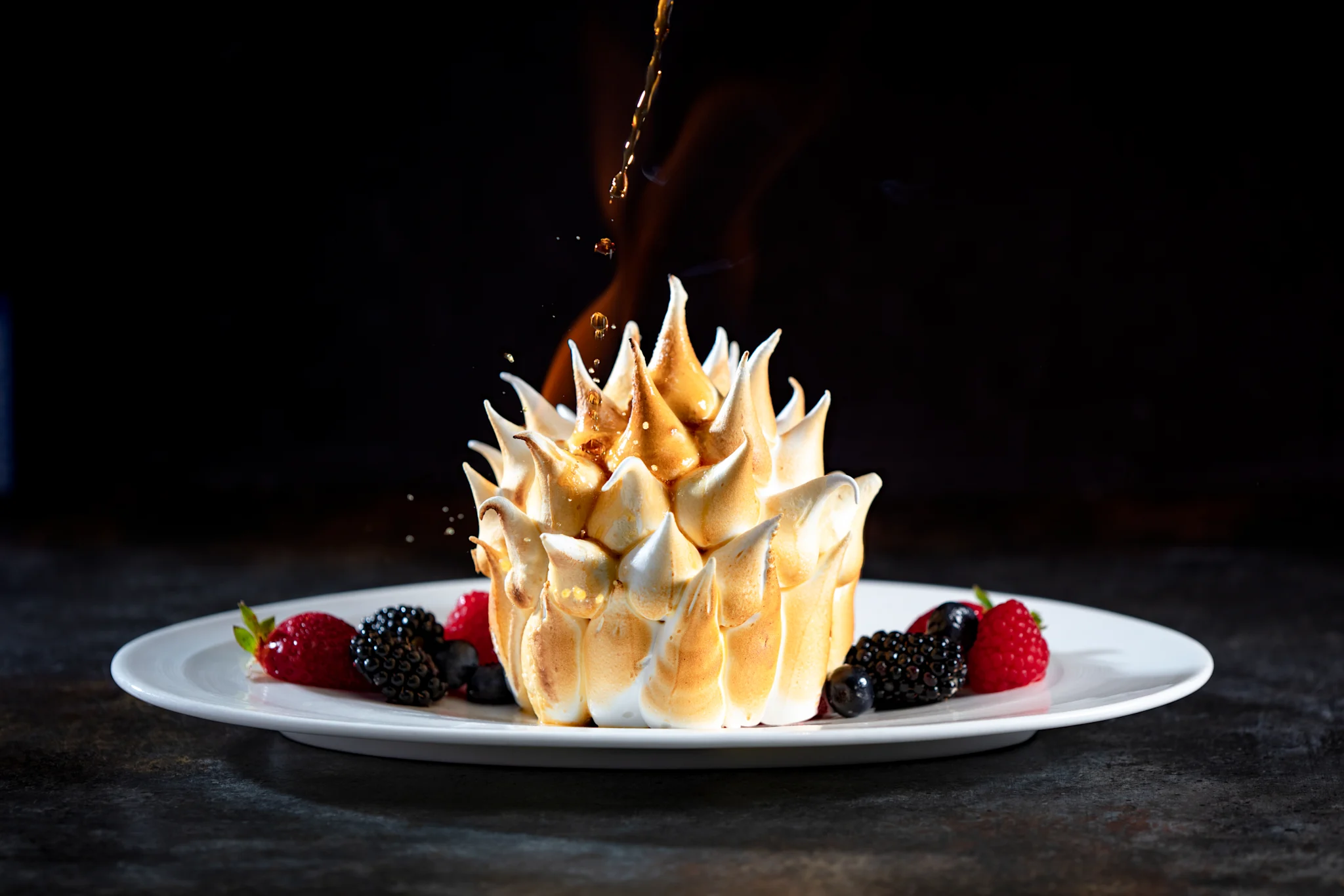 Toasted meringue dessert with fresh berries on white plate, caramel being drizzled on top.