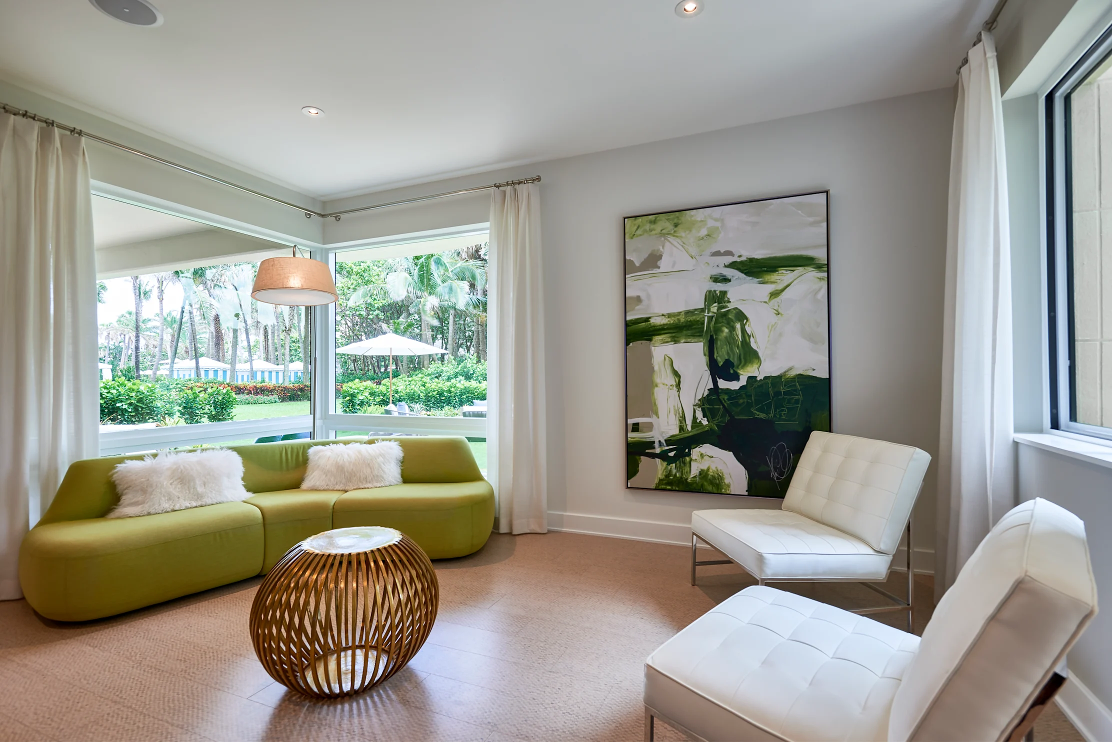 Modern living room with green curved sofa, white chairs, botanical artwork, and garden view window.