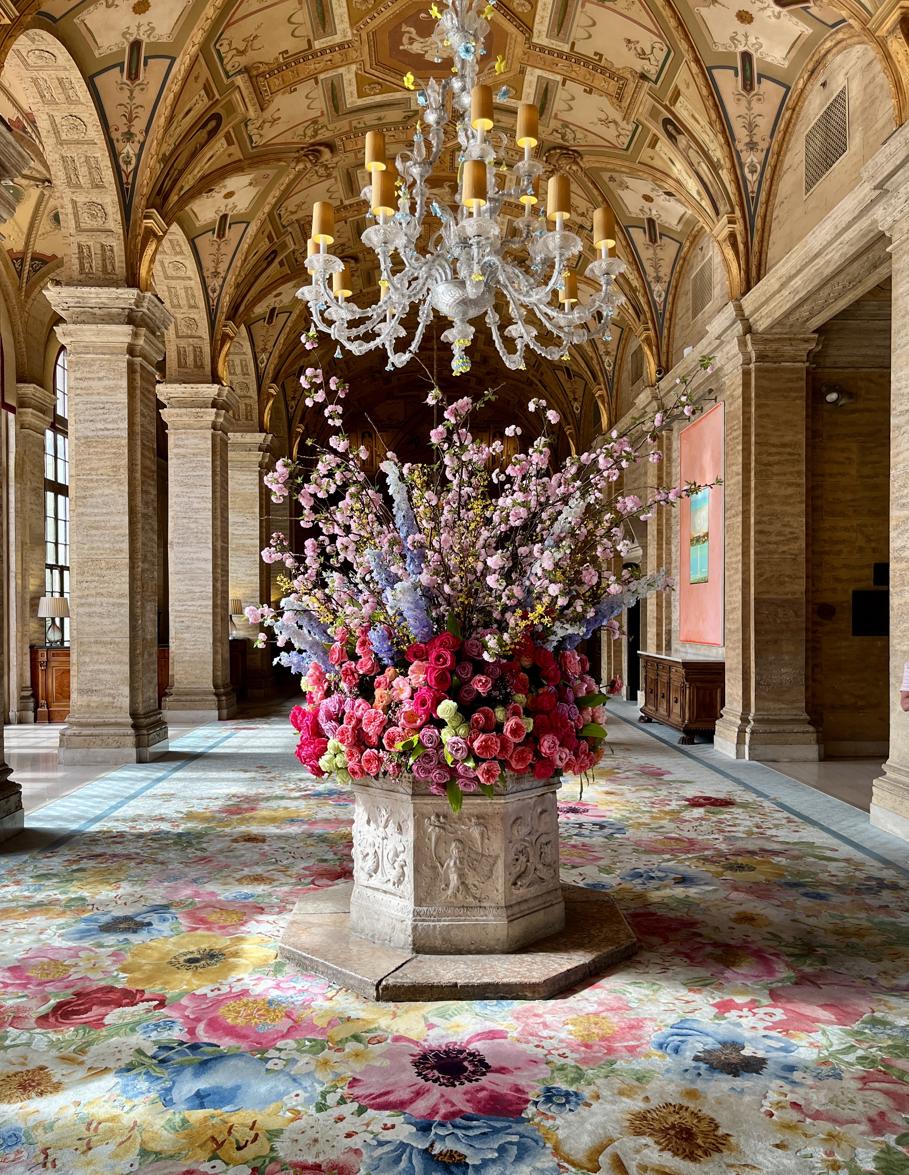 Ornate floral arrangement in historic vaulted hall with crystal chandelier and decorated ceiling.