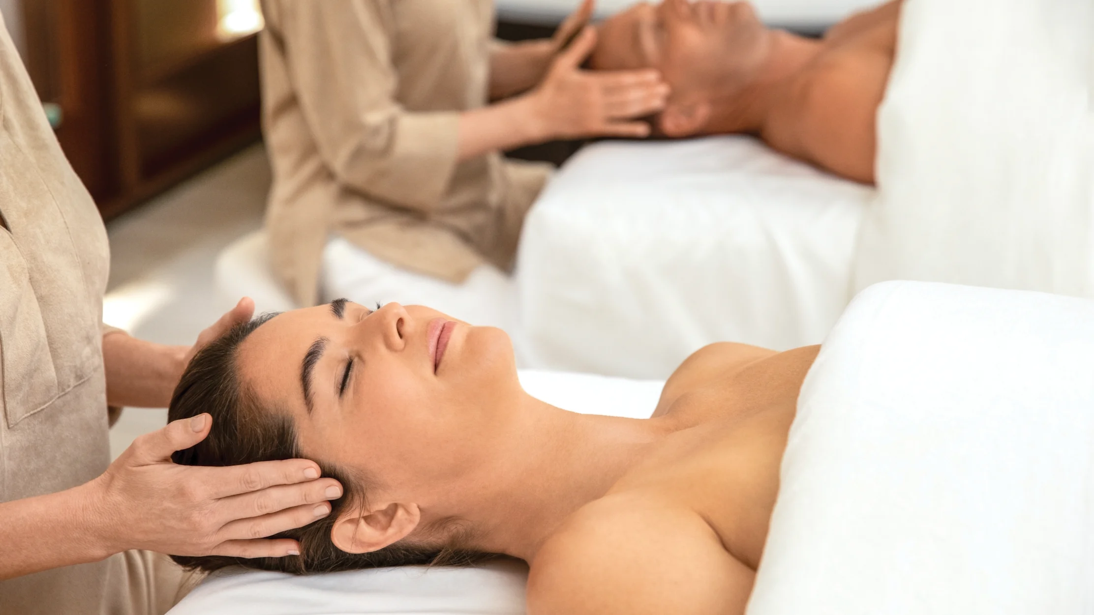Two people receiving relaxing head massages at a spa, lying on white massage tables.