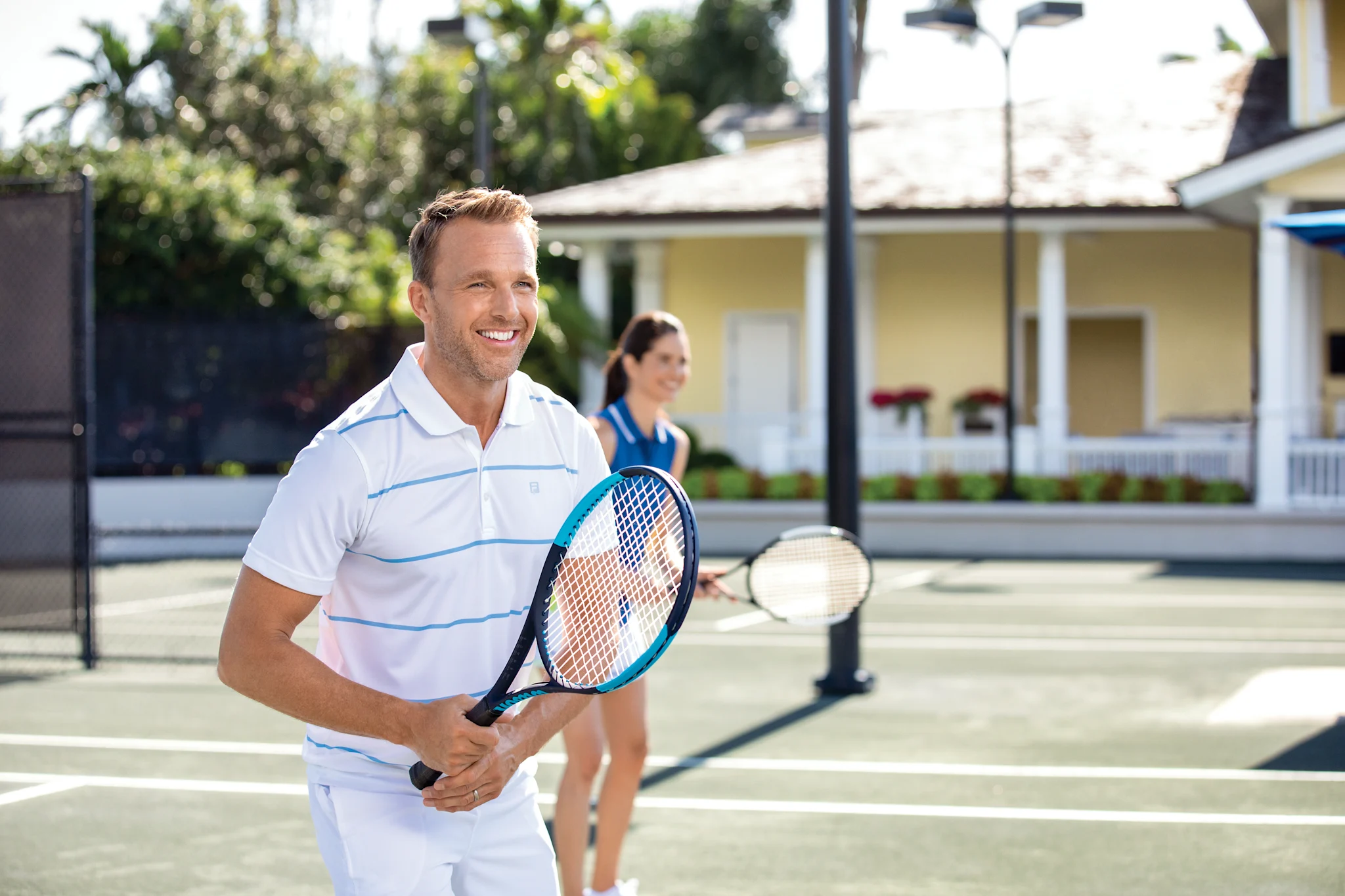 Smiling man in white striped polo holding tennis racket on outdoor court with woman in background.