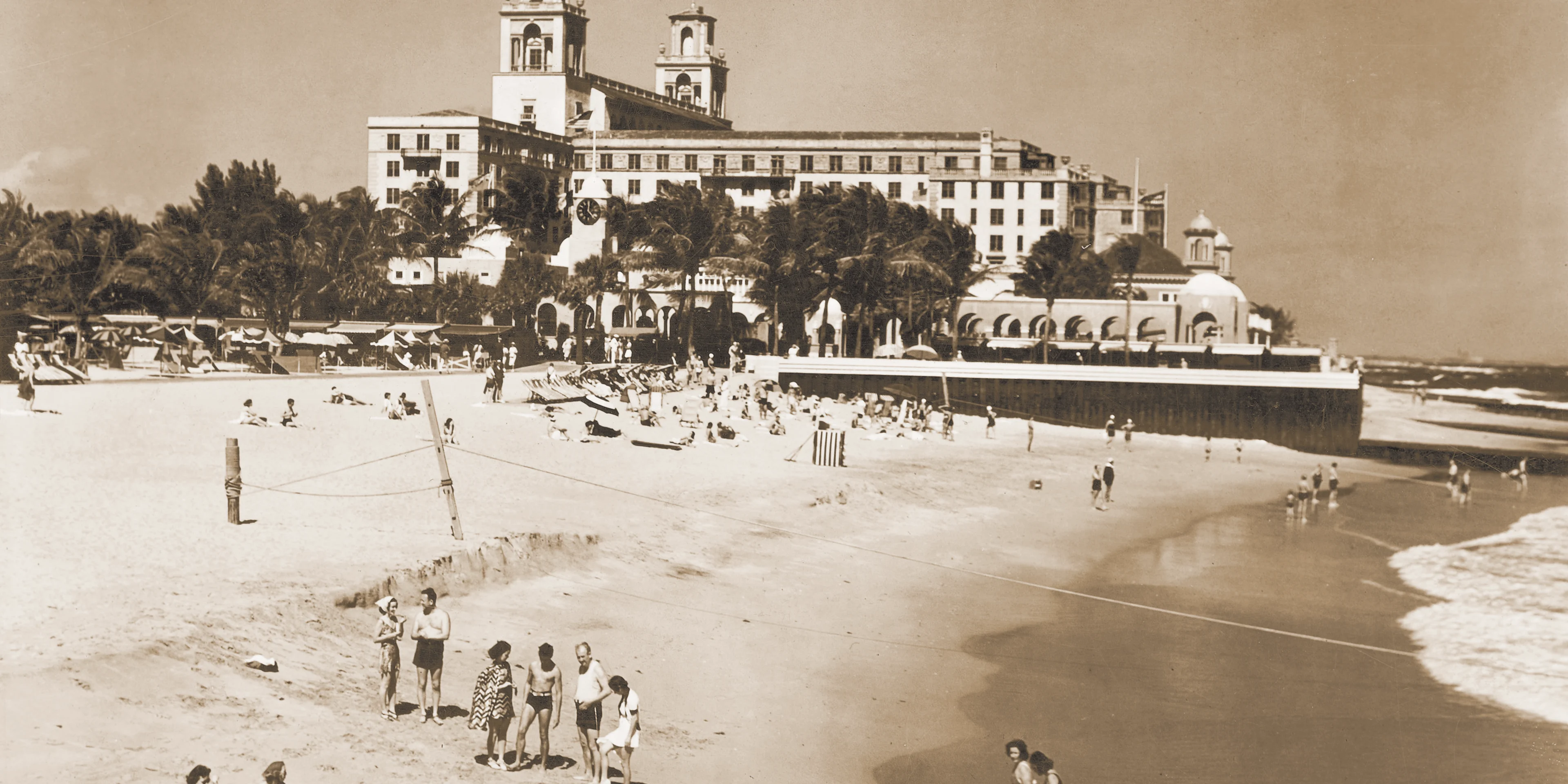 Vintage sepia photograph of The Breakers Palm beach behind beachgoers on sand with a large white Spanish-style hotel in background.