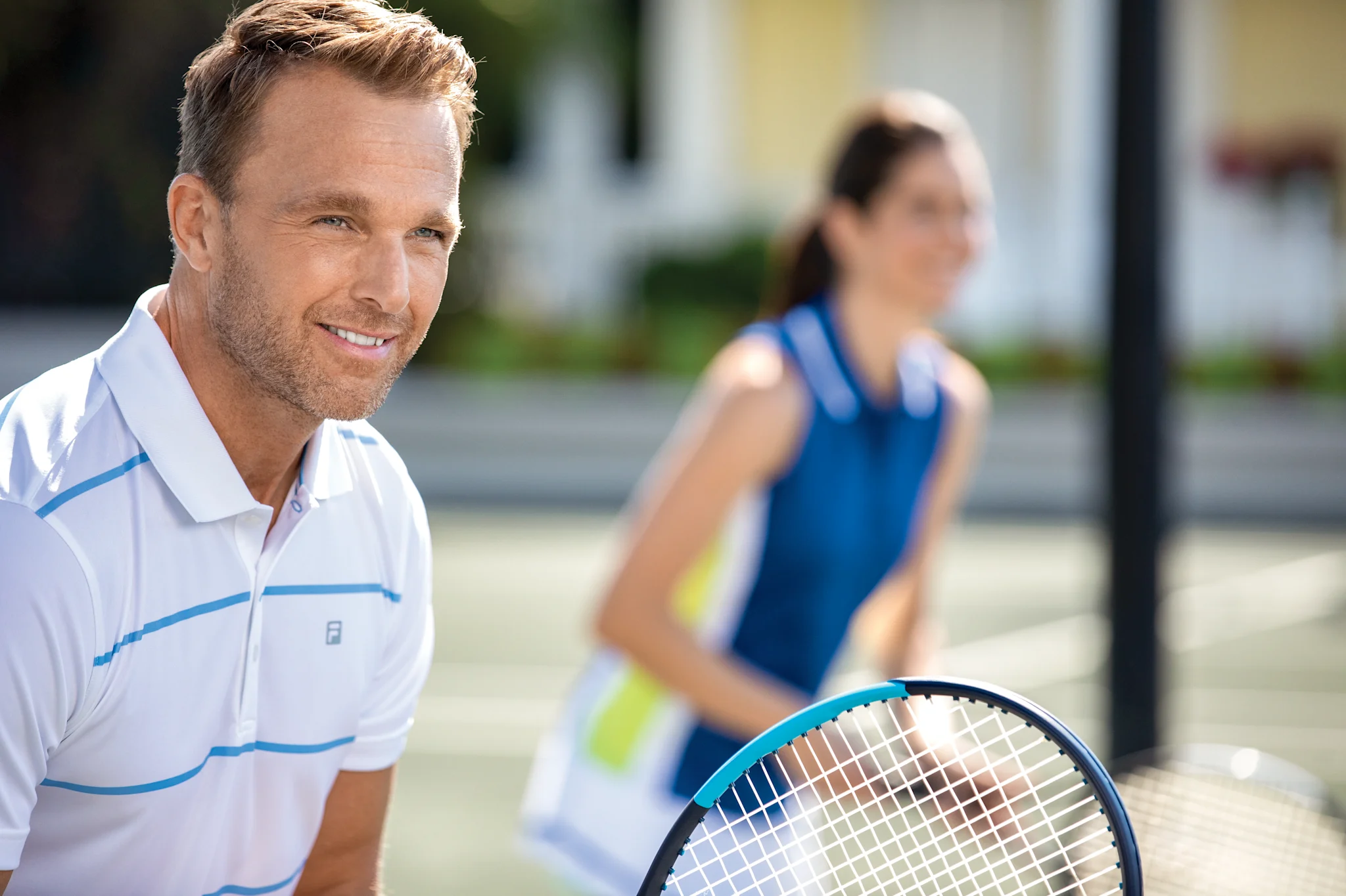 Smiling man in white tennis shirt holding blue racket with woman player in background on court