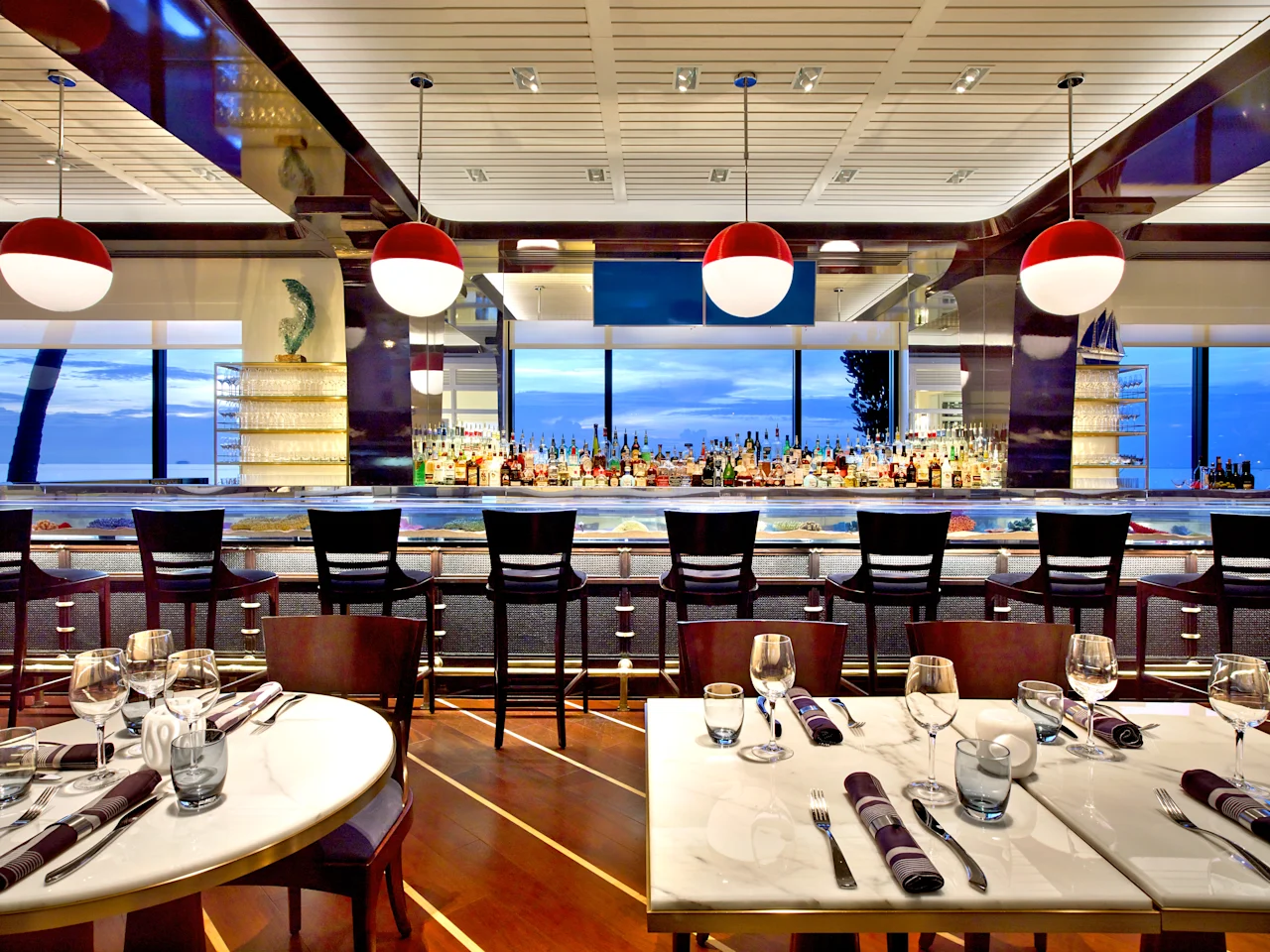 Modern restaurant interior with red pendant lights, bar with liquor bottles, ocean view windows, and tables.