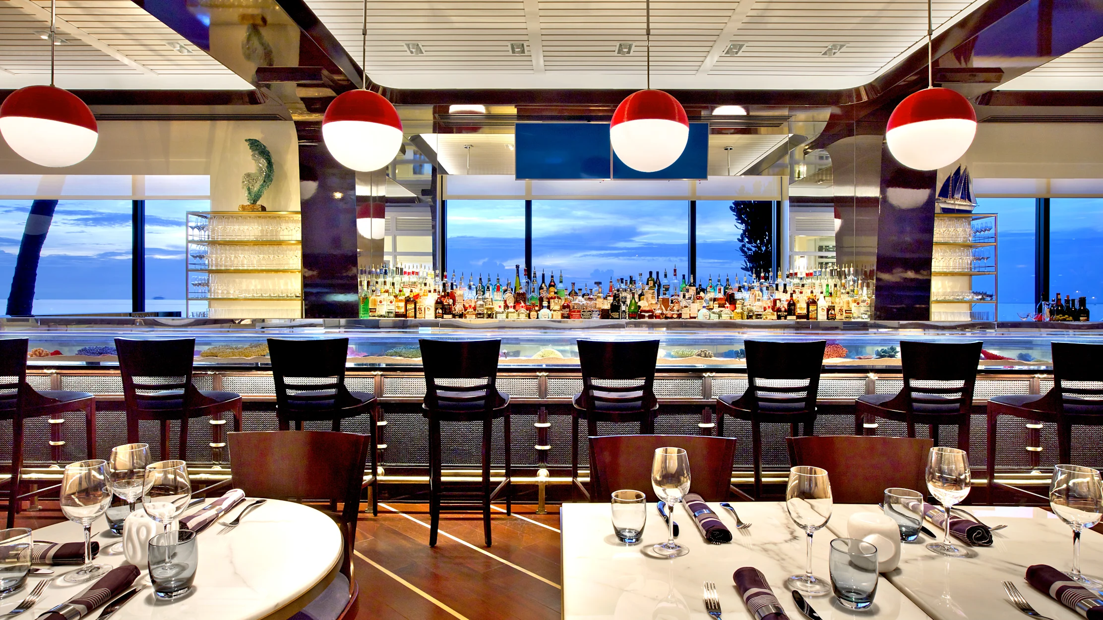 Modern restaurant interior with red pendant lights, bar with liquor bottles, ocean view windows, and tables.