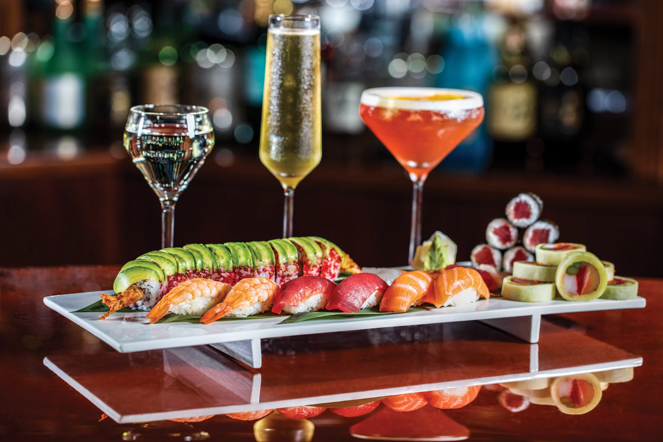 Assorted sushi platter with three cocktails on a bar counter with blurred bottles in background.
