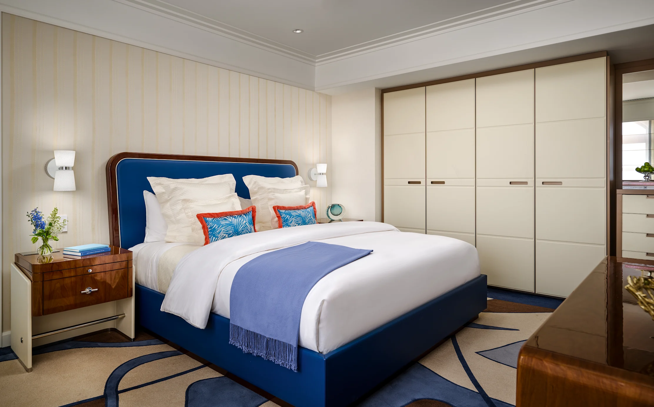 Modern bedroom with blue upholstered bed, white linens, cream wardrobes, and wood accents.