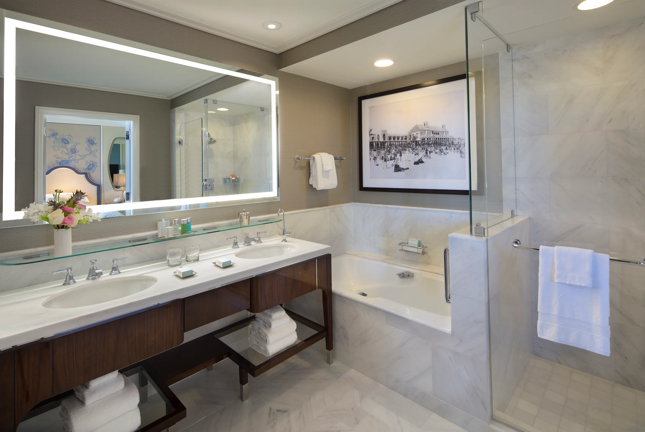 Modern hotel bathroom with double vanity, backlit mirror, soaking tub, glass shower, and vintage beach photo.