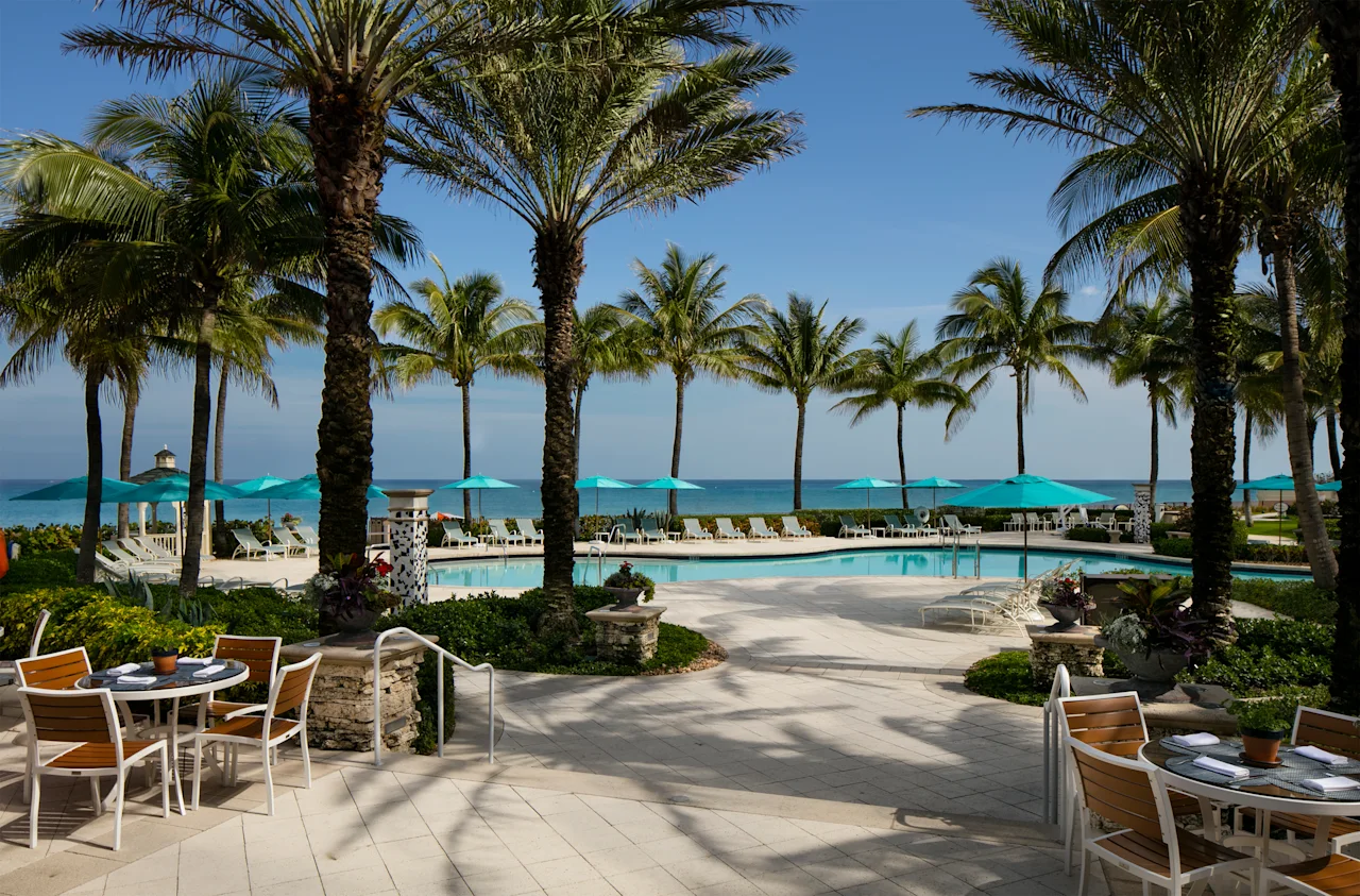 Tropical resort pool area with palm trees, turquoise umbrellas, lounge chairs, and ocean view.