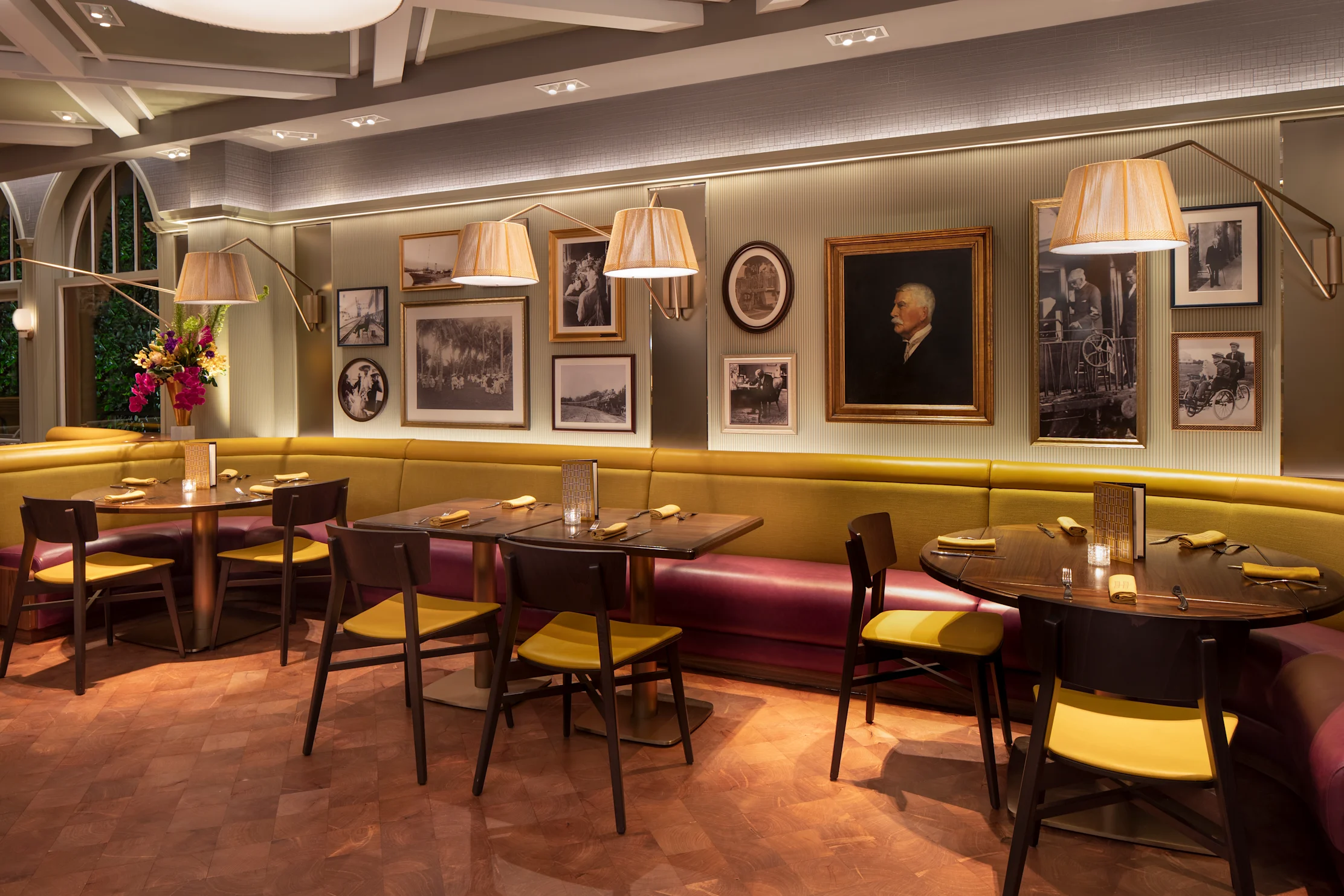 Elegant restaurant interior with yellow banquette seating, vintage portraits on gray walls, and arc lamps.