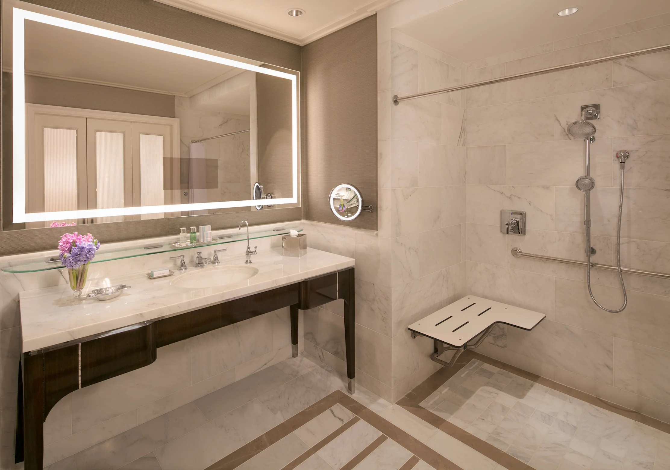 Modern accessible bathroom with marble vanity, backlit mirror, and roll-in shower with bench seat.