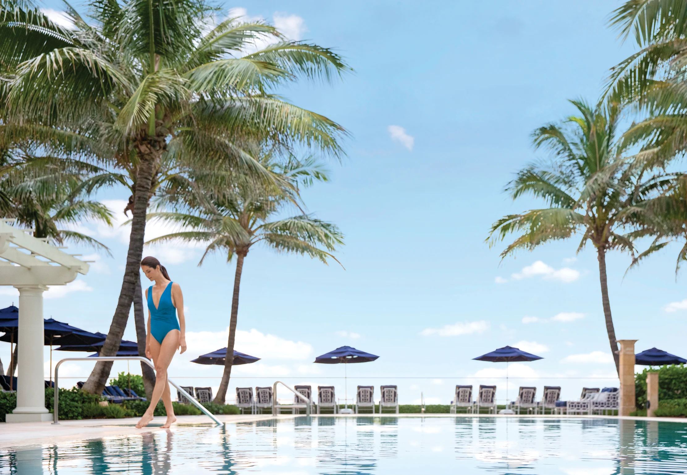 Woman in blue swimsuit walking by tropical resort pool with palm trees and lounge chairs under sunny sky.