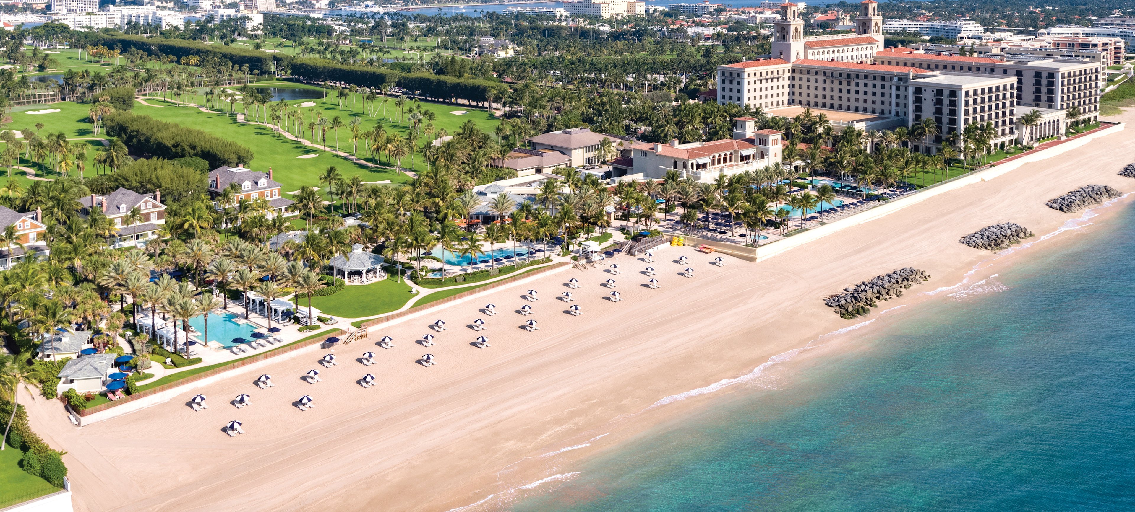 Aerial view of beachfront resort with white sand, turquoise water, golf course, and palm trees.