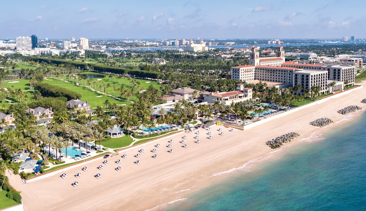 Aerial view of beachfront resort with white sand, turquoise water, golf course, and palm trees.