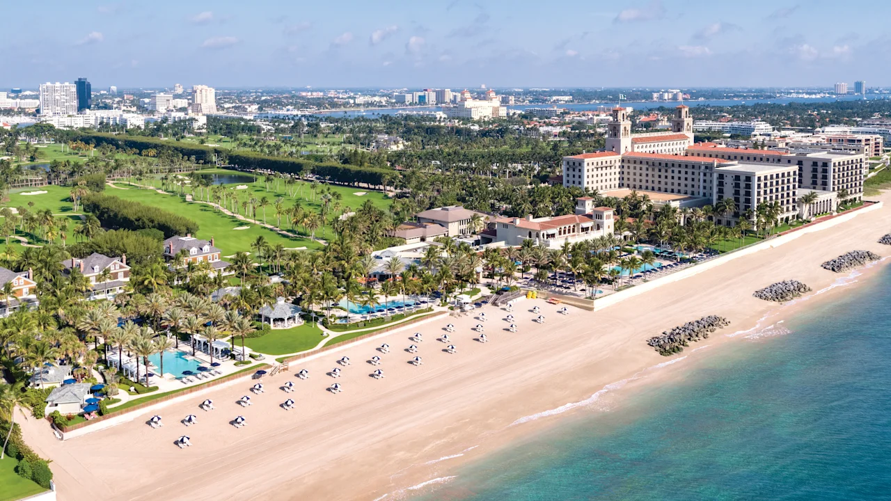 Aerial view of beachfront resort with white sand, turquoise water, golf course, and palm trees.