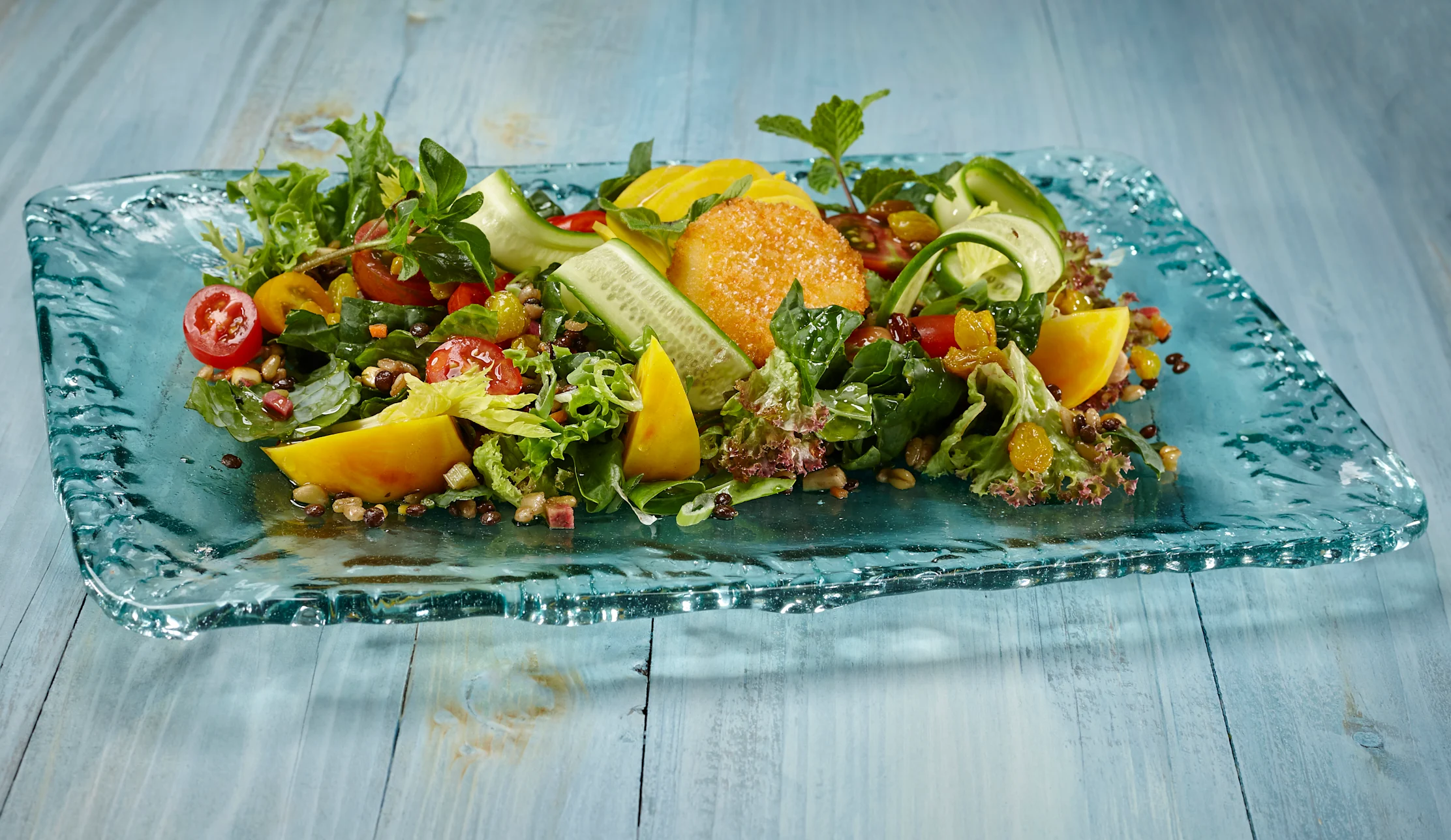 Colorful salad with greens, tomatoes, yellow peppers, cucumber ribbons, and quinoa on blue glass plate.