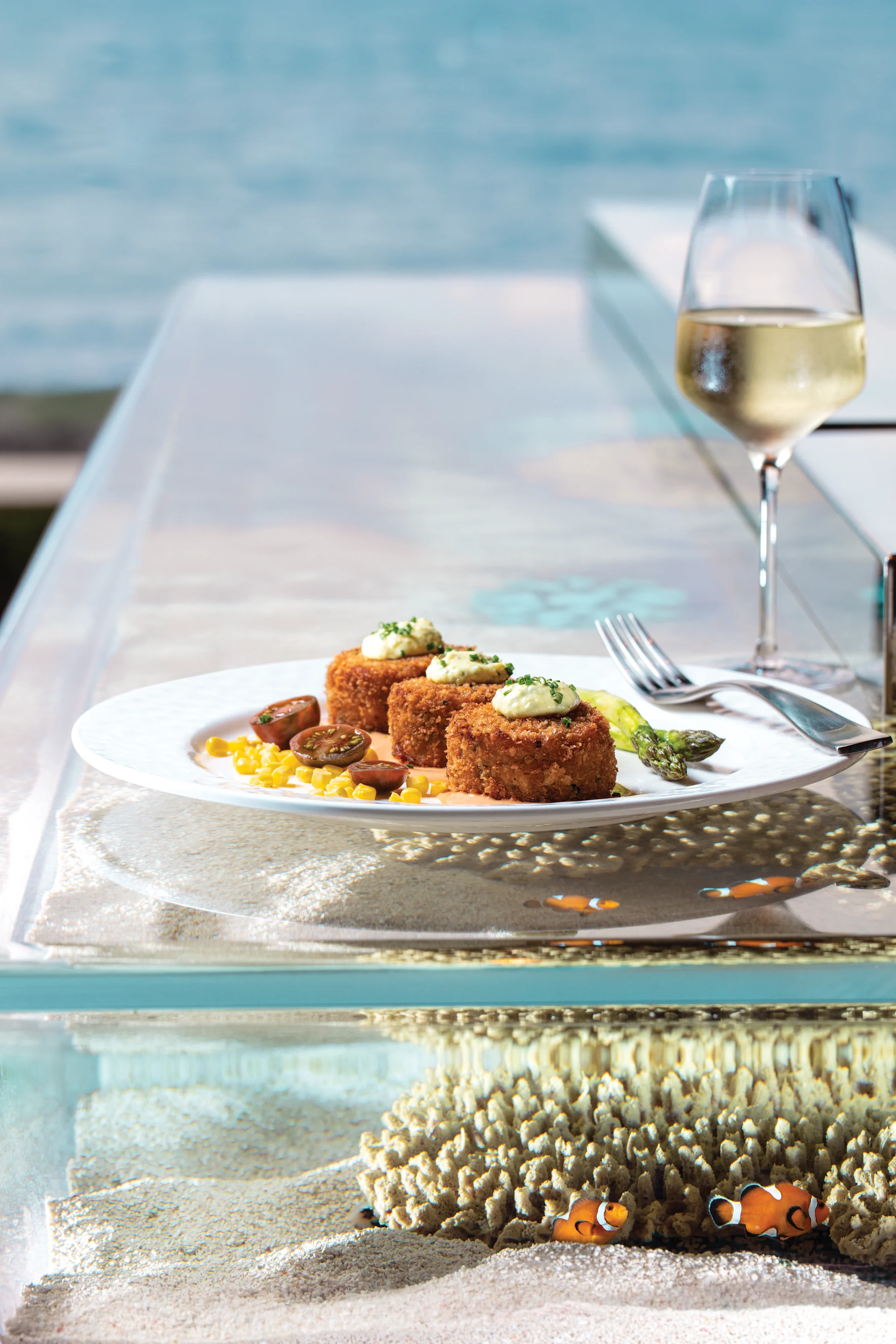 Seafood dish with breaded crab cakes and vegetables on white plate beside white wine glass.
