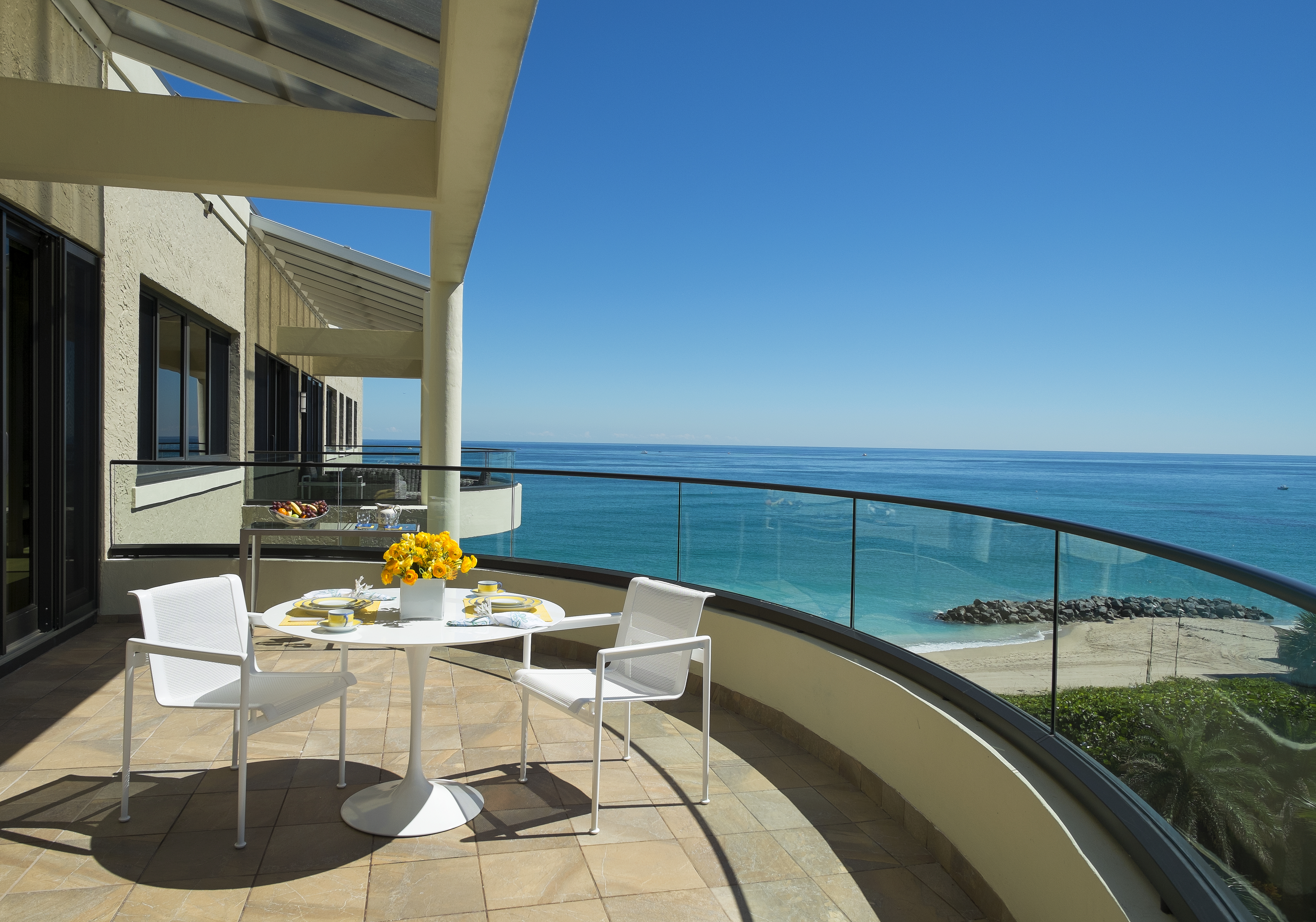 Oceanfront balcony with white dining set, yellow flowers, and glass railing overlooking turquoise water.