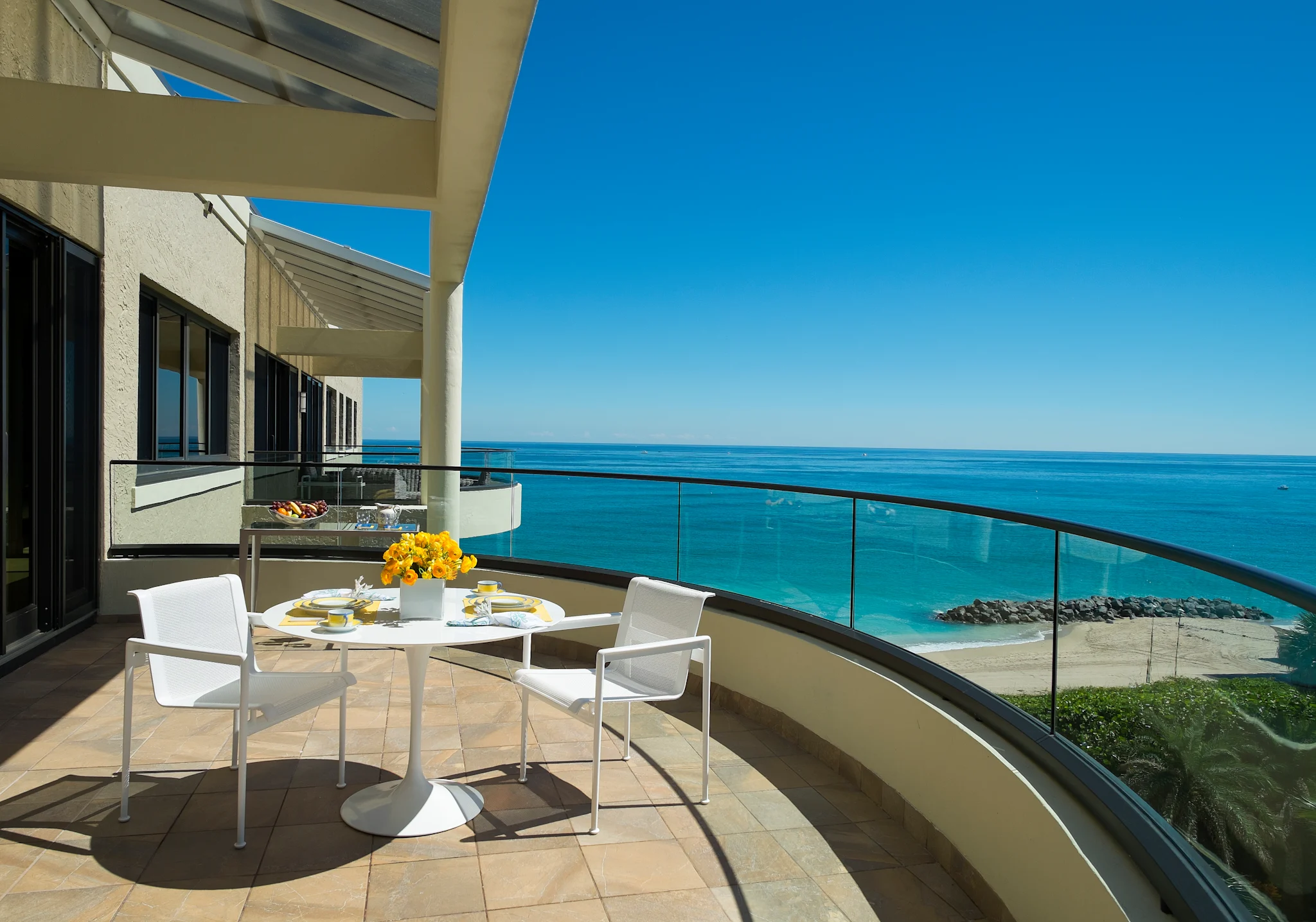 Oceanfront balcony with white dining set, yellow flowers, and glass railing overlooking turquoise water.