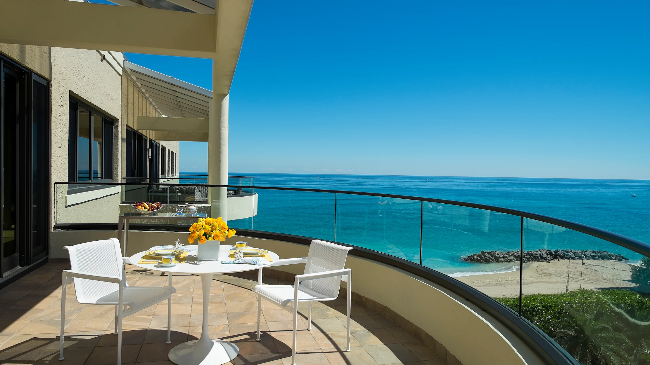 Oceanfront balcony with white dining set, yellow flowers, and glass railing overlooking turquoise water.