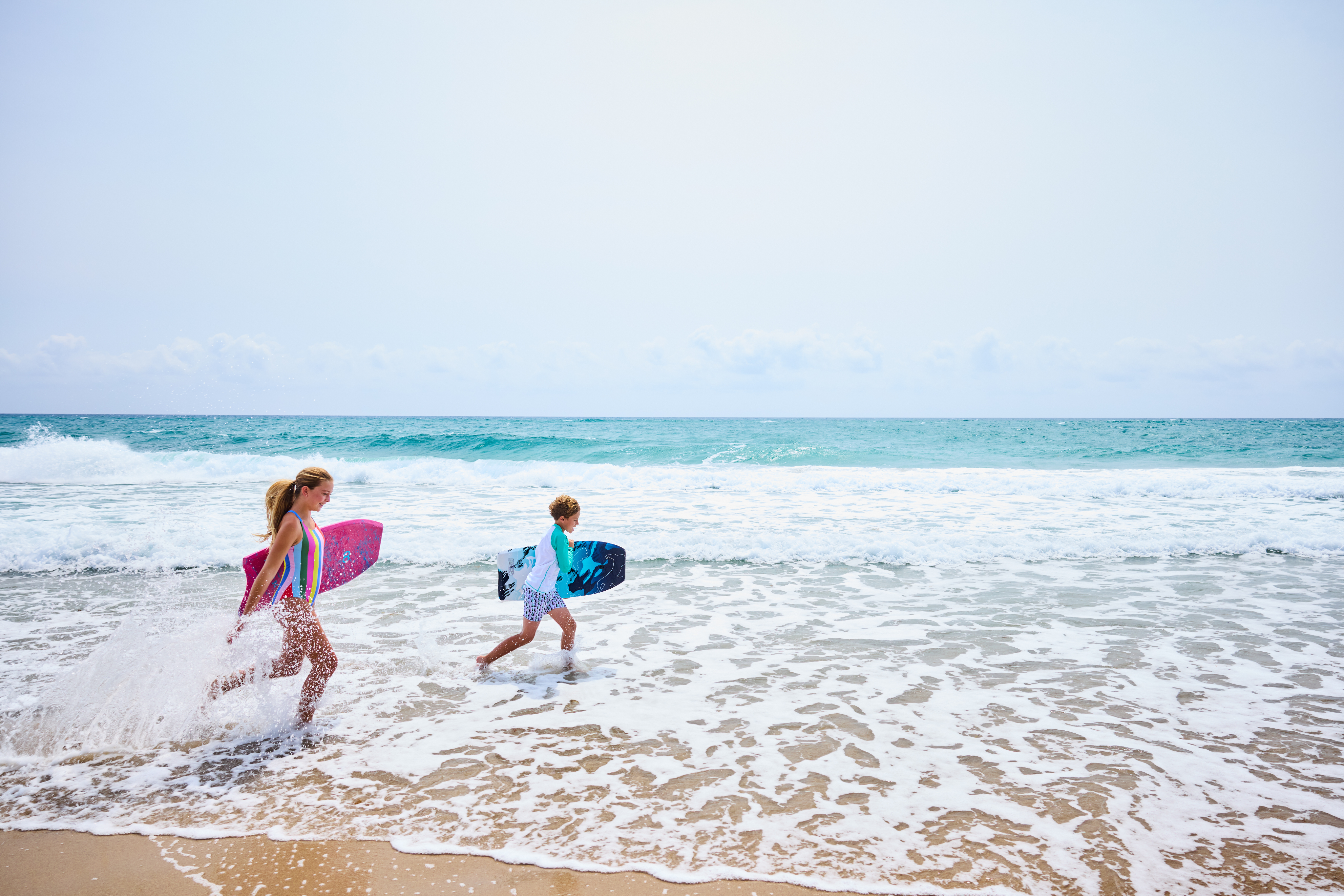 Two children running through ocean waves carrying colorful boogie boards on a sunny beach day.
