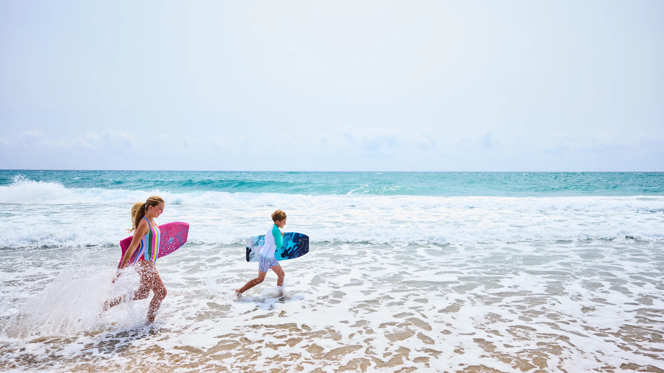 Two children running through ocean waves carrying colorful boogie boards on a sunny beach day.