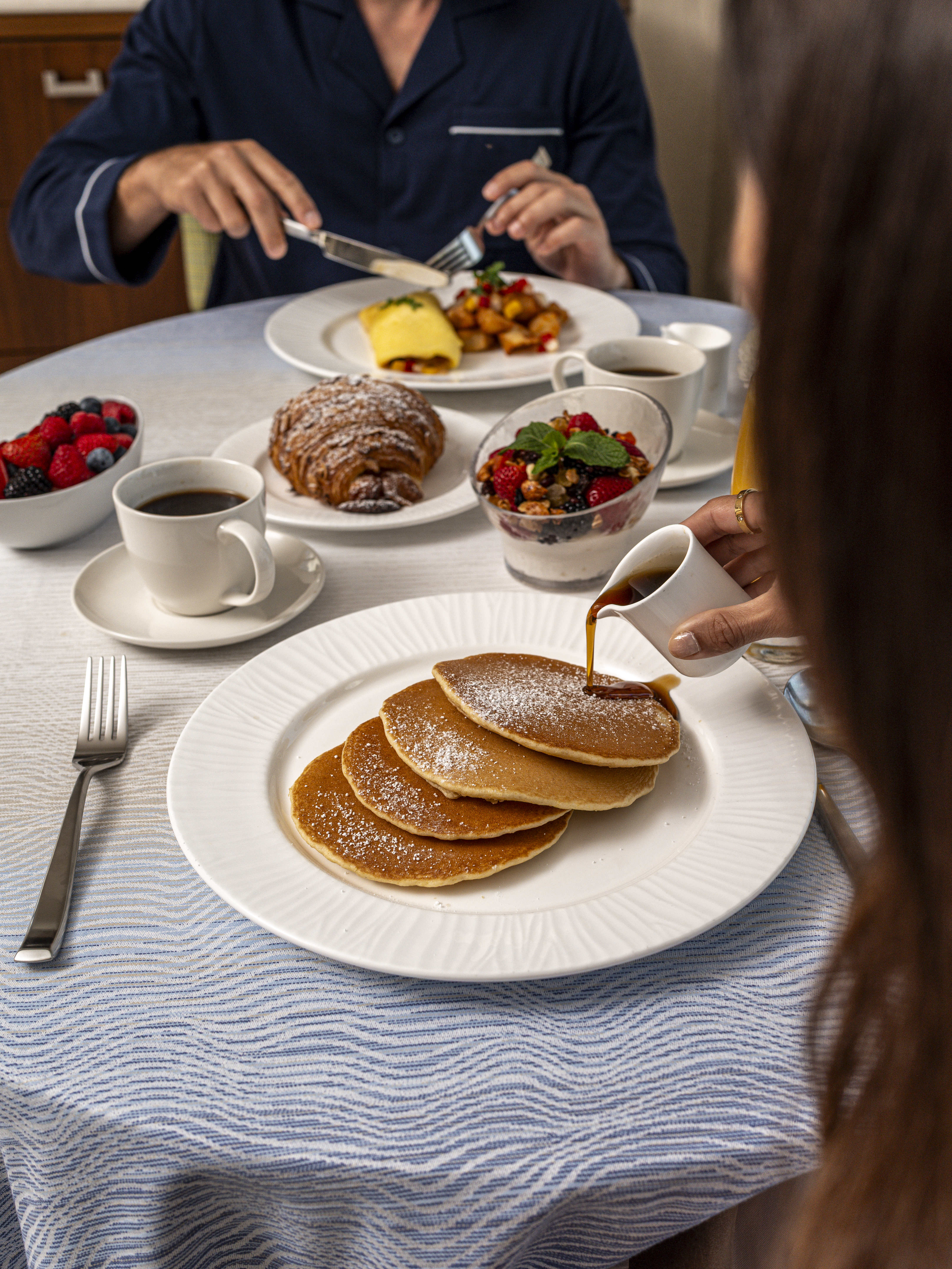 Hand pouring syrup over stack of powdered sugar pancakes with breakfast spread including coffee and fruit.