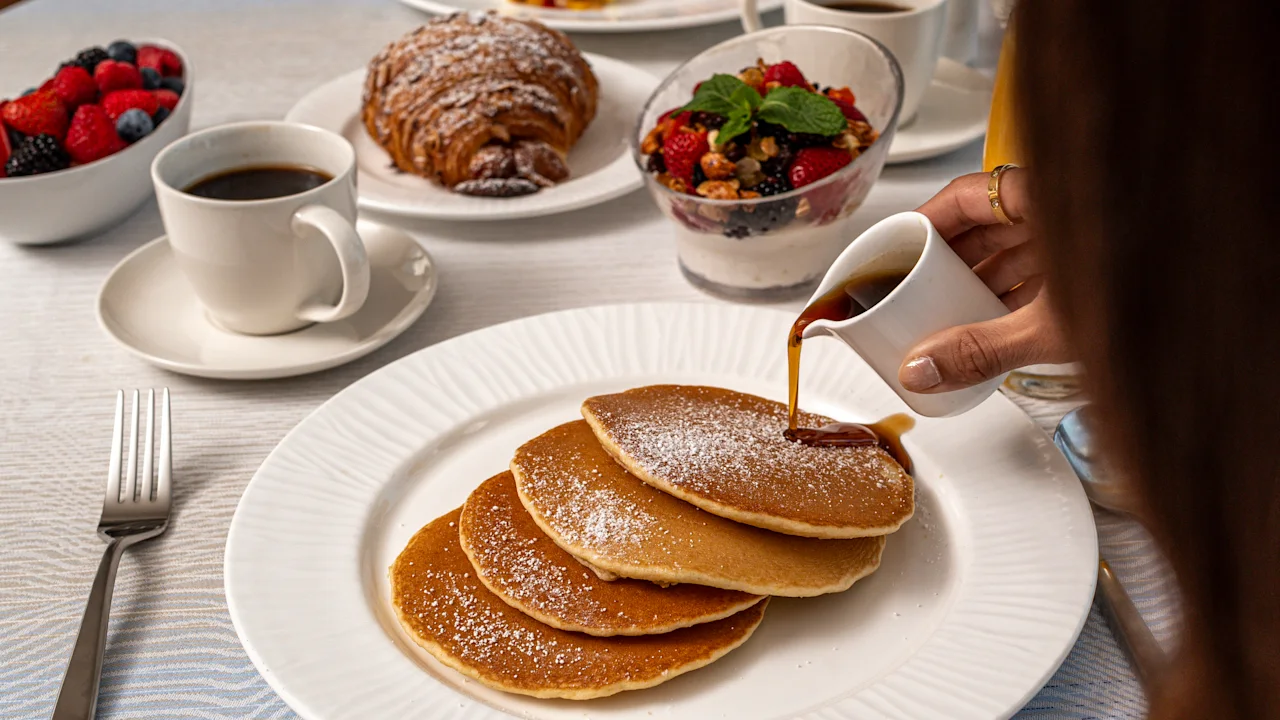 Hand pouring syrup over stack of powdered sugar pancakes with breakfast spread including coffee and fruit.