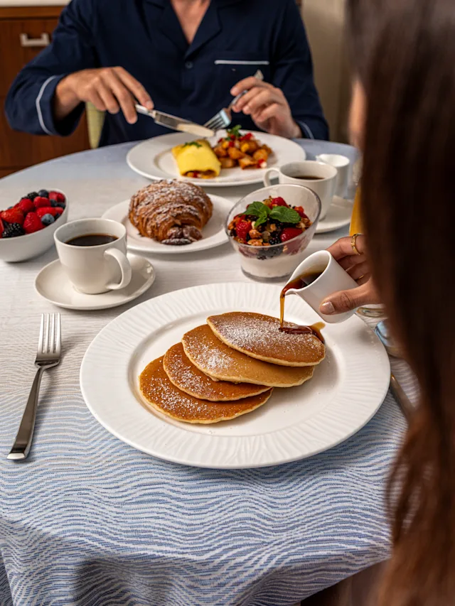 Hand pouring syrup over stack of powdered sugar pancakes with breakfast spread including coffee and fruit.
