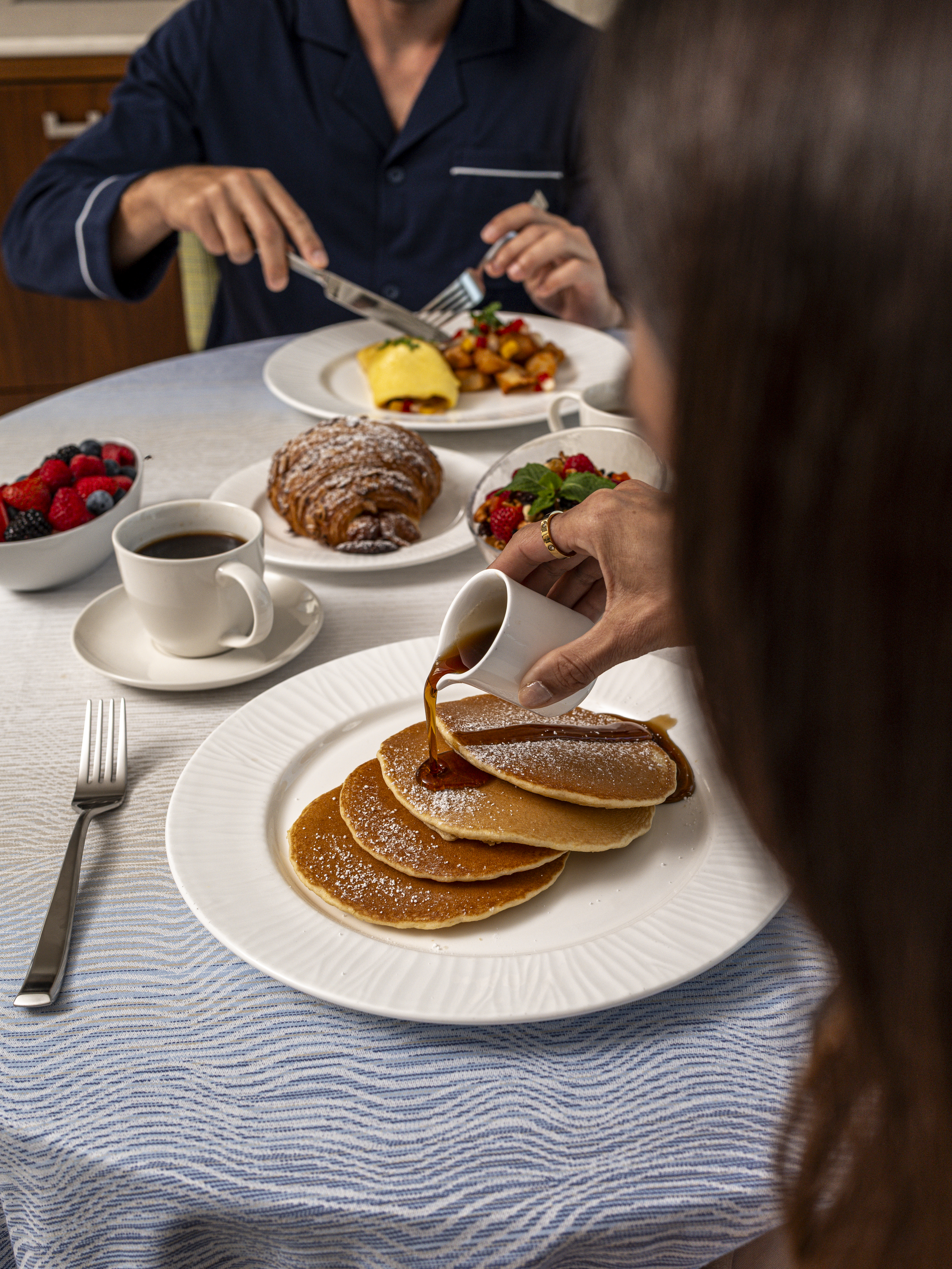 Hand pouring syrup over stack of powdered sugar pancakes with breakfast spread including coffee and fruit.