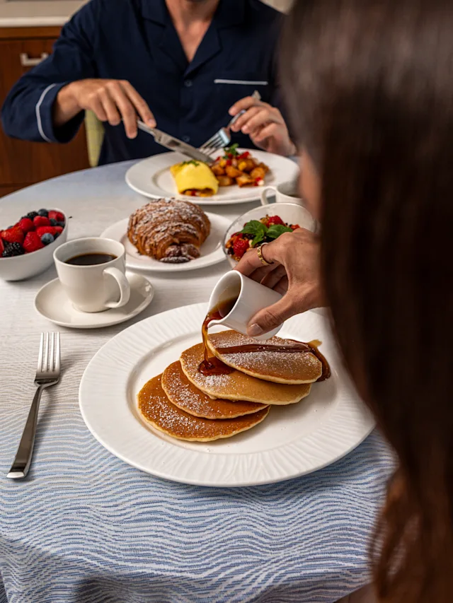 Hand pouring syrup over stack of powdered sugar pancakes with breakfast spread including coffee and fruit.