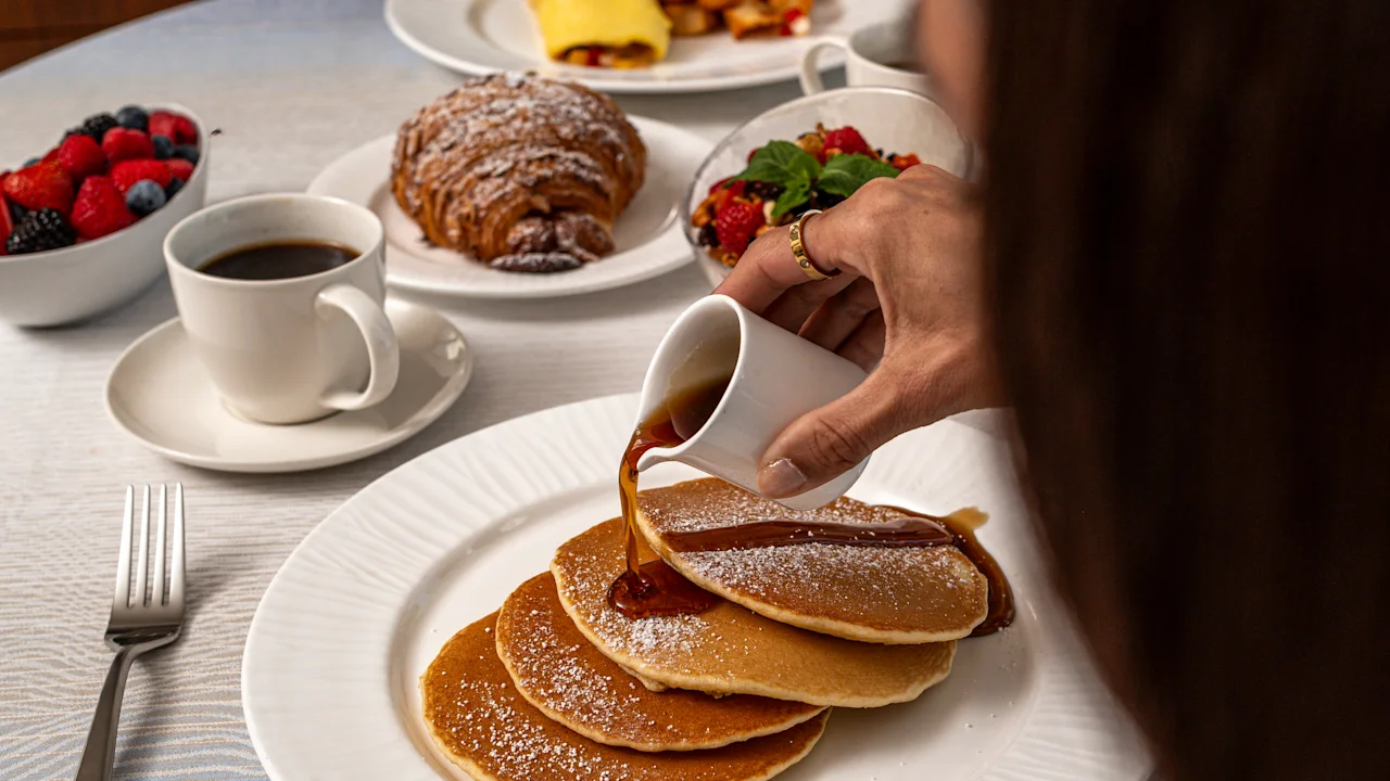 Hand pouring syrup over stack of powdered sugar pancakes with breakfast spread including coffee and fruit.