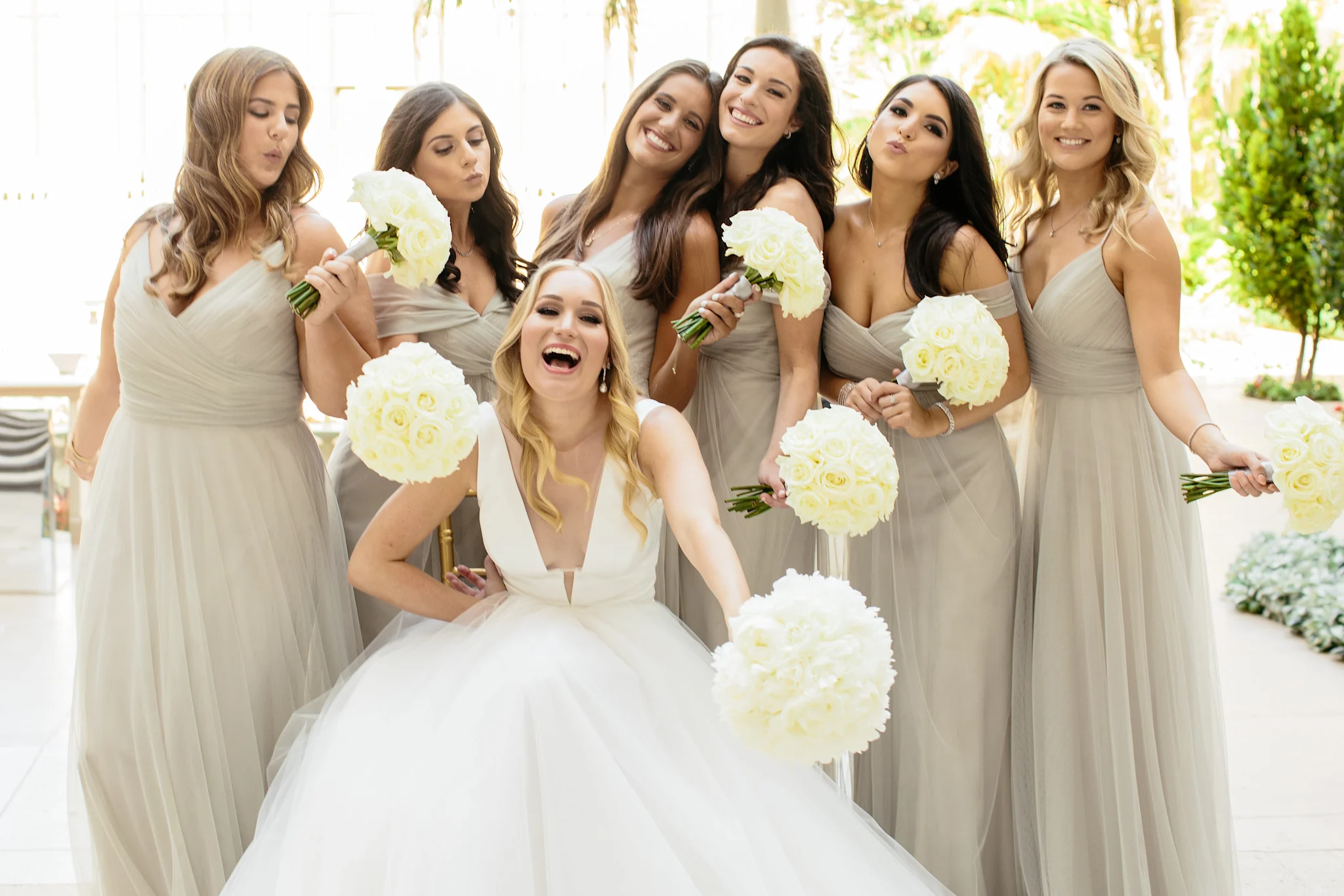 Bride in white gown with six bridesmaids in beige dresses holding white rose bouquets outdoors.