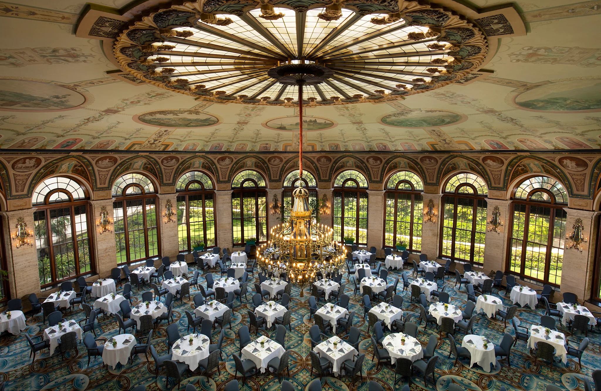 Ornate ballroom with painted ceiling dome, large chandelier, arched windows, and round tables set for dining.