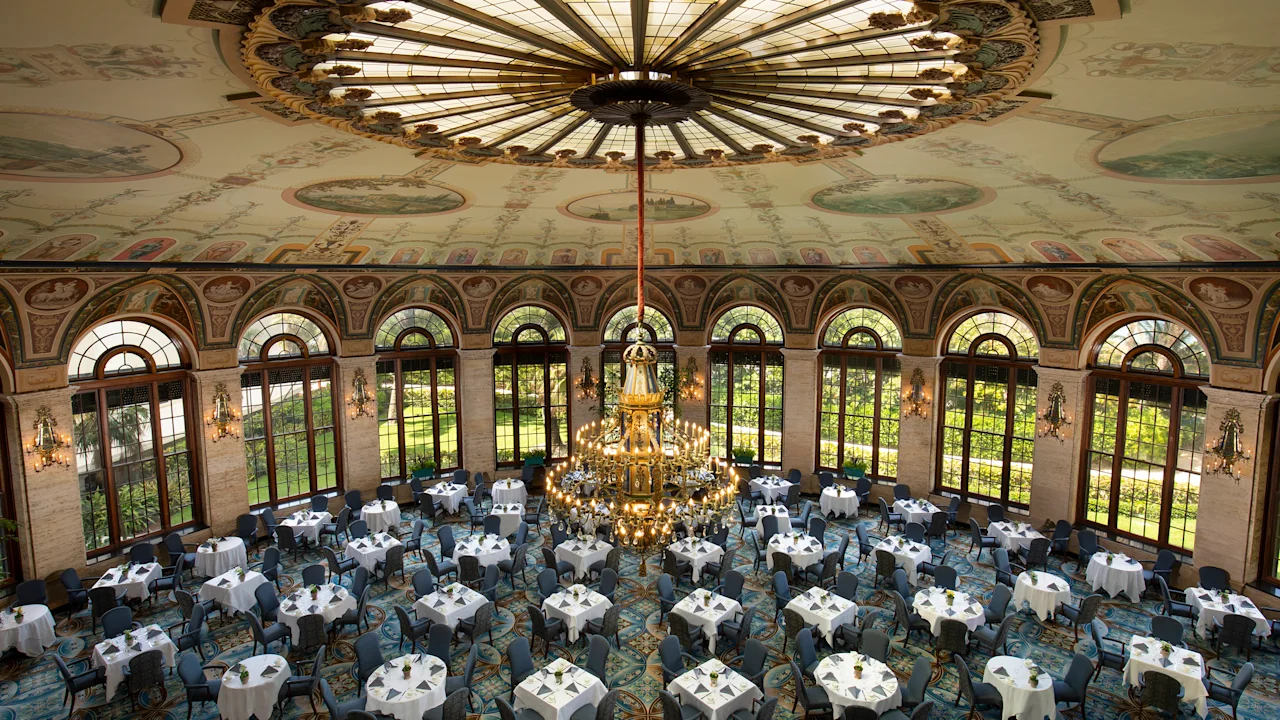 Ornate ballroom with painted ceiling dome, large chandelier, arched windows, and round tables set for dining.