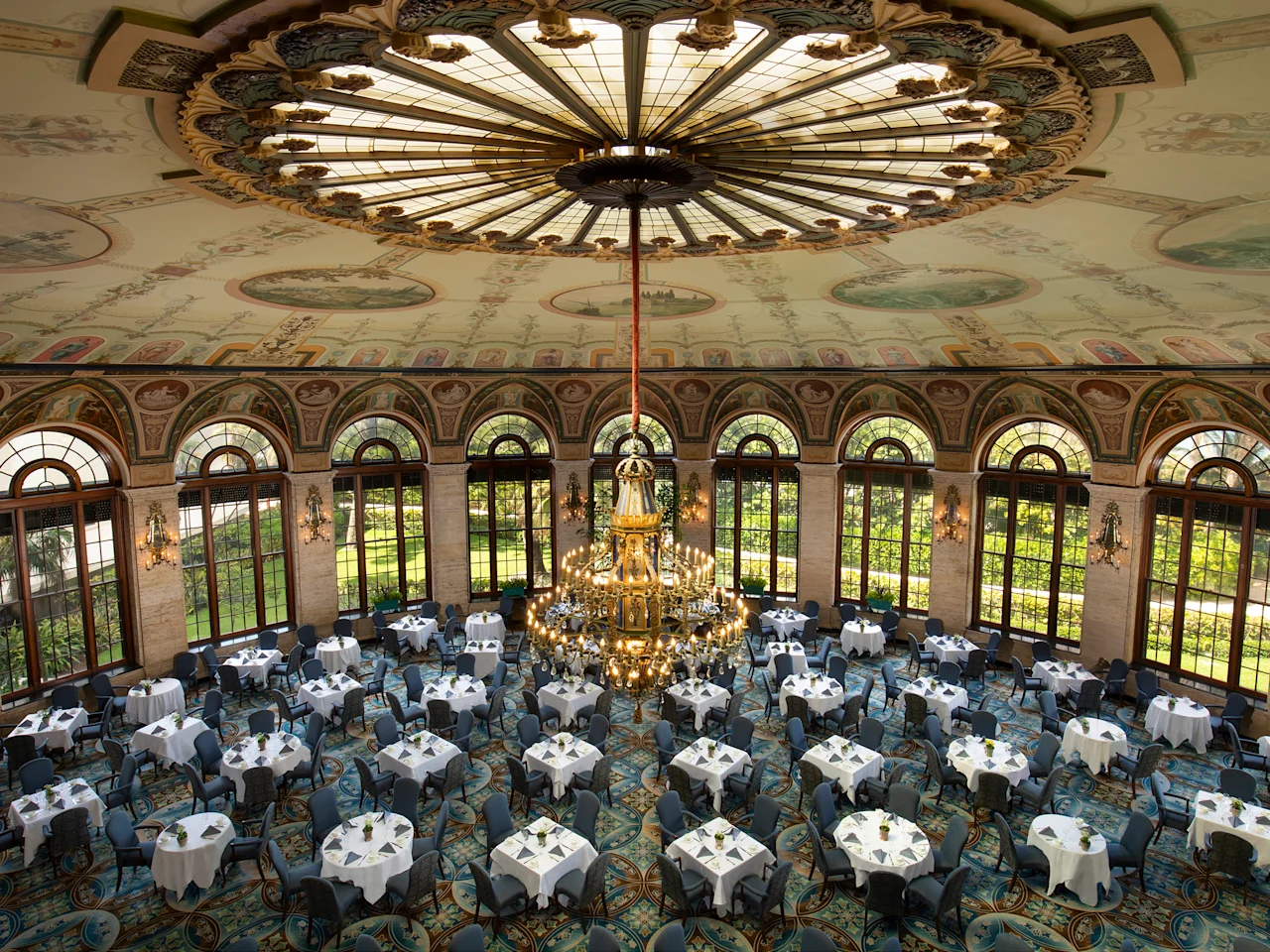 Ornate ballroom with painted ceiling dome, large chandelier, arched windows, and round tables set for dining.