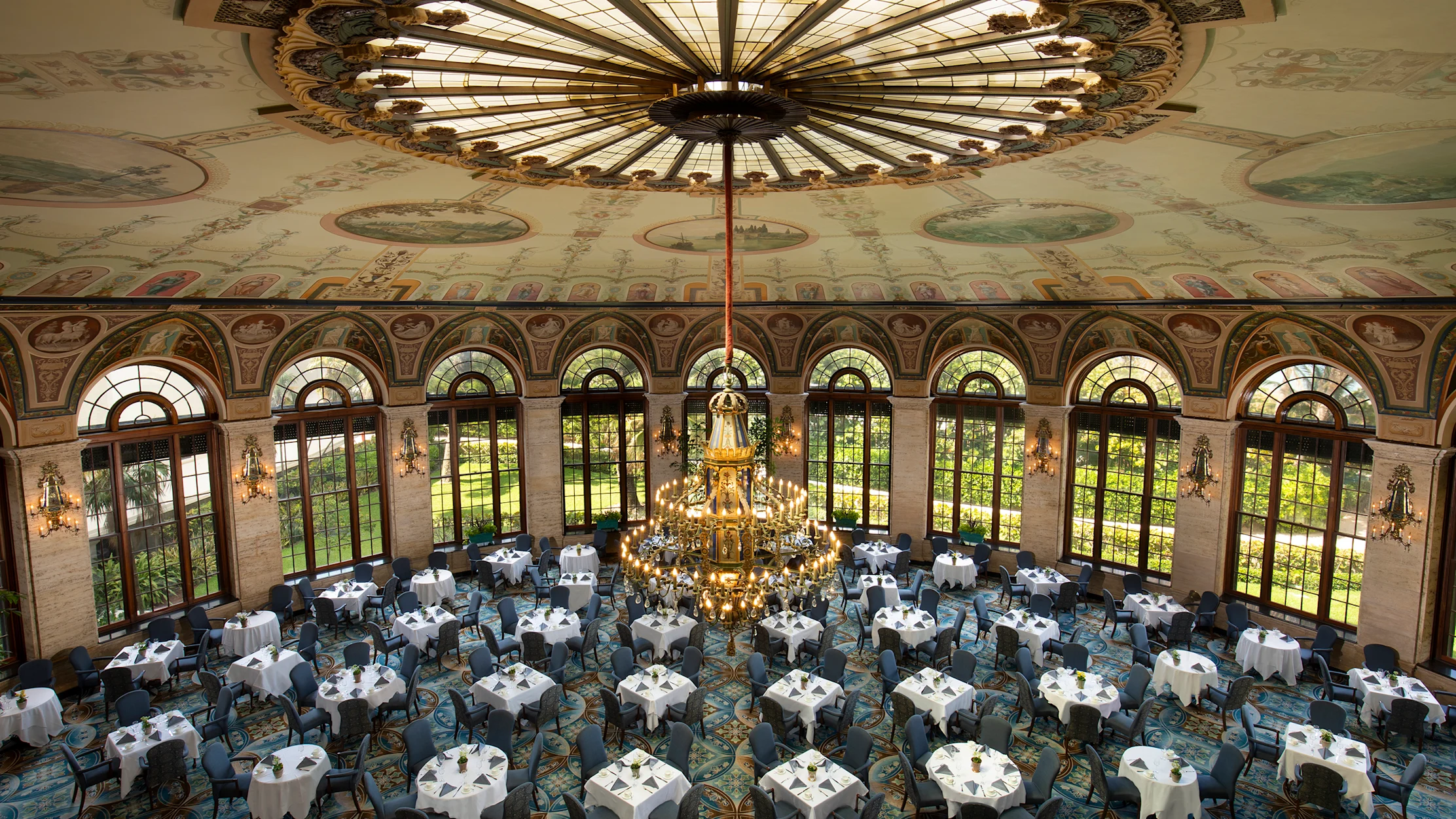 Ornate ballroom with painted ceiling dome, large chandelier, arched windows, and round tables set for dining.