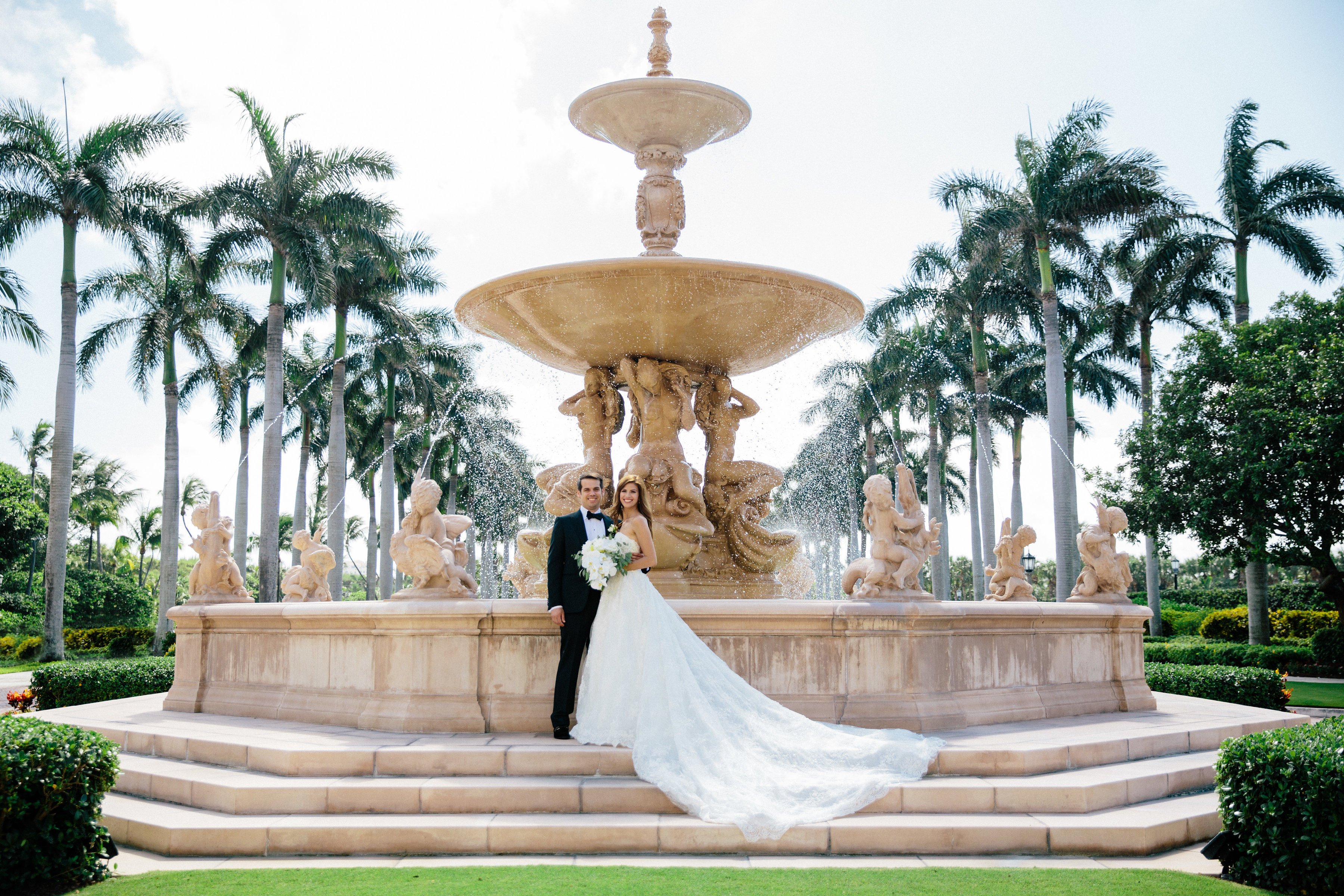 Bride and groom pose in front of ornate stone fountain surrounded by palm trees in tropical setting.