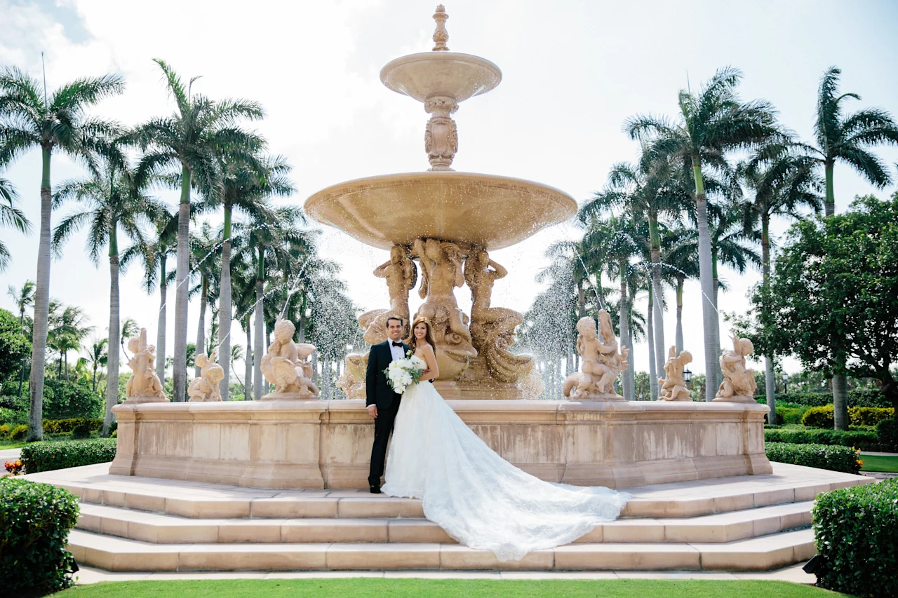 Bride and groom pose in front of ornate stone fountain surrounded by palm trees in tropical setting.