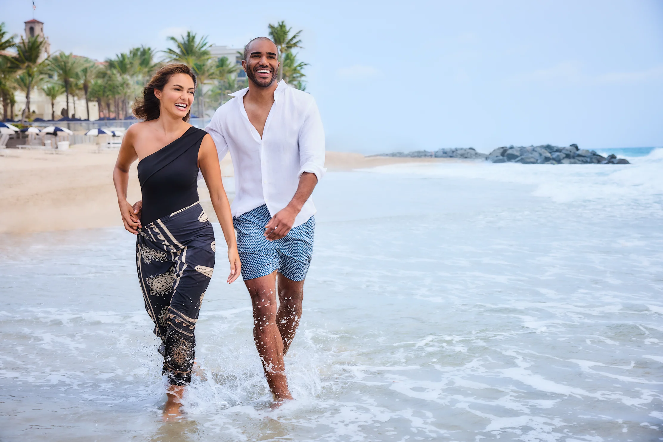 Happy couple walking hand in hand through shallow ocean waves on a tropical beach with palm trees.