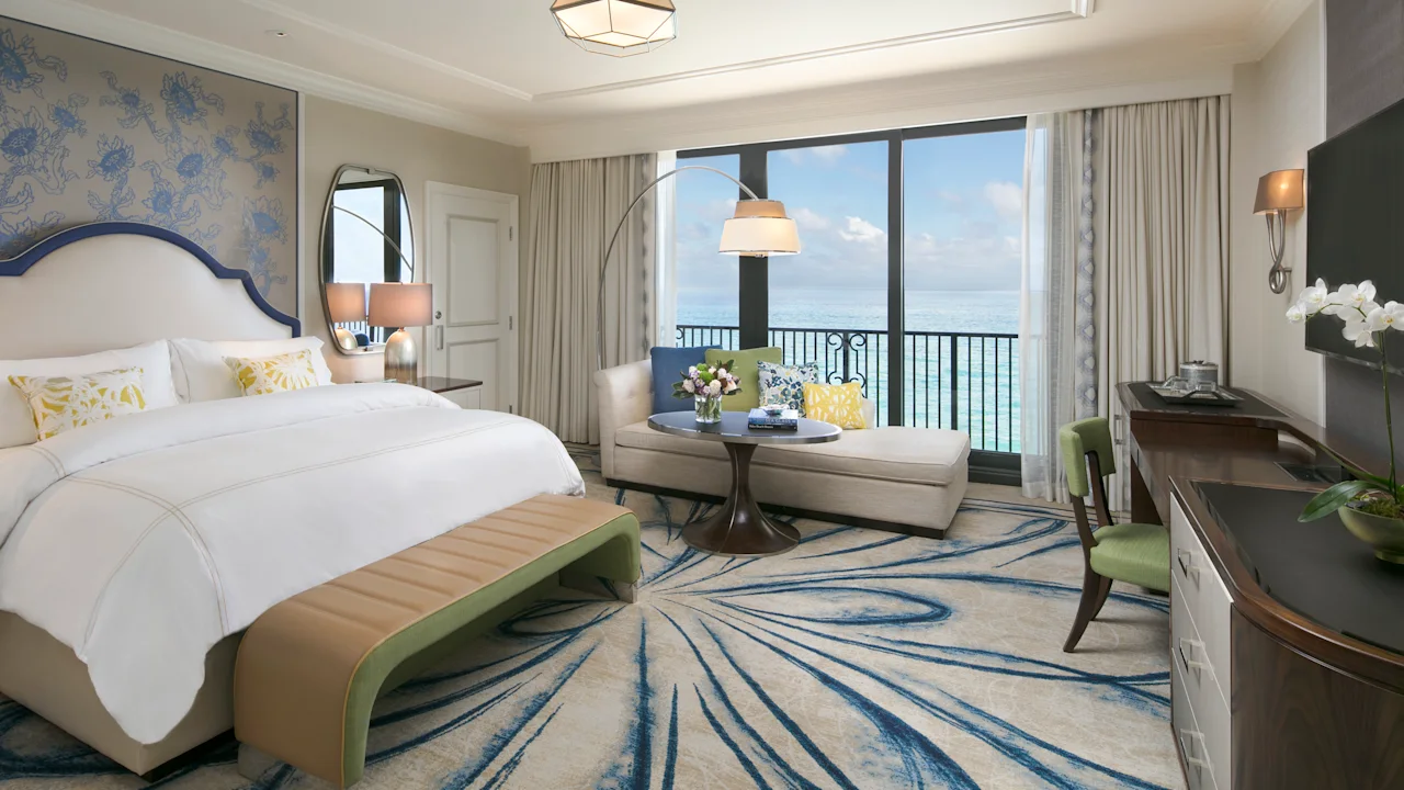 Elegant hotel room with ocean view, white bed, blue accents, seating area, and balcony overlooking water.