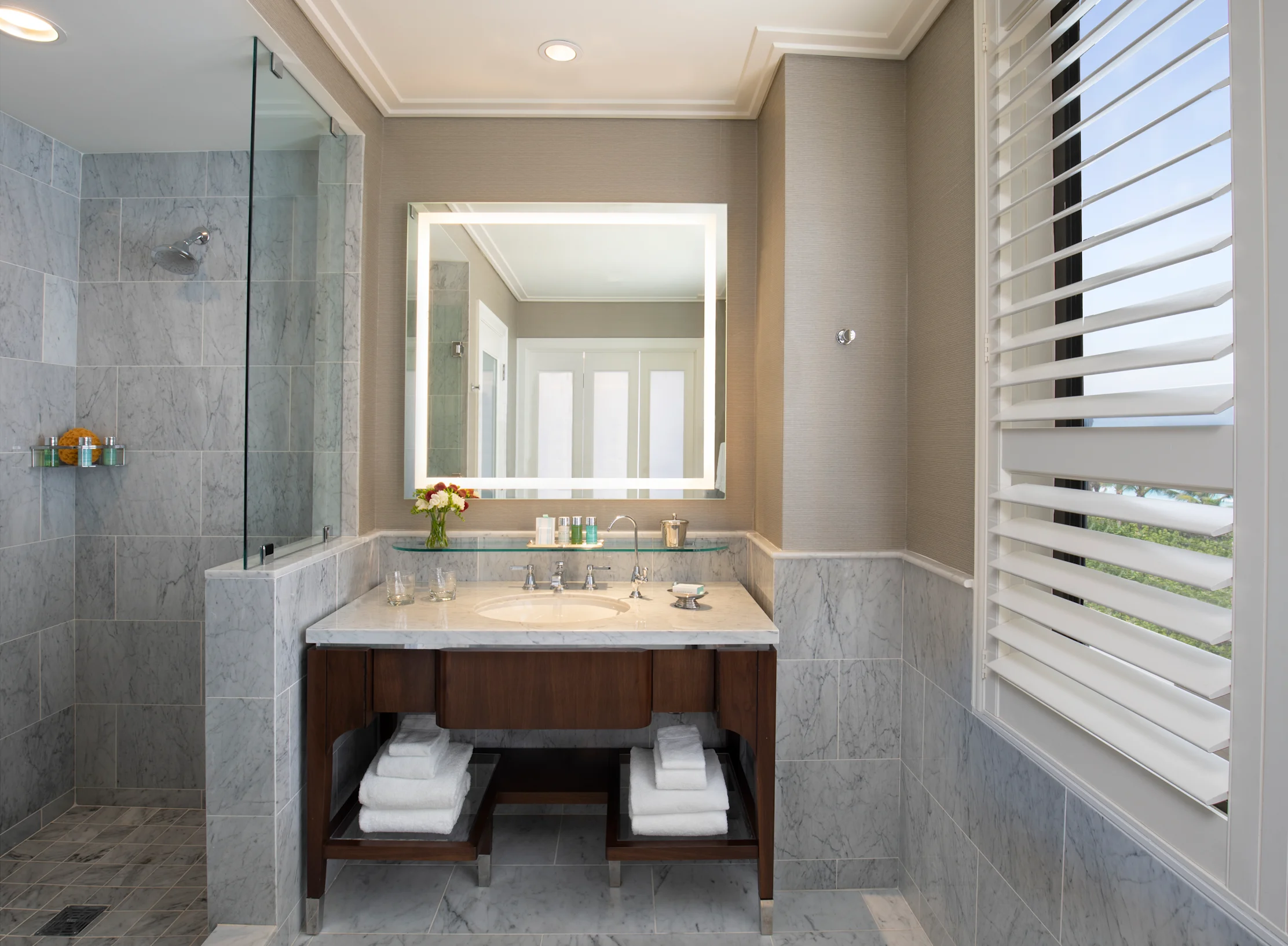 Modern bathroom with marble vanity, illuminated mirror, glass shower, and white plantation shutters.