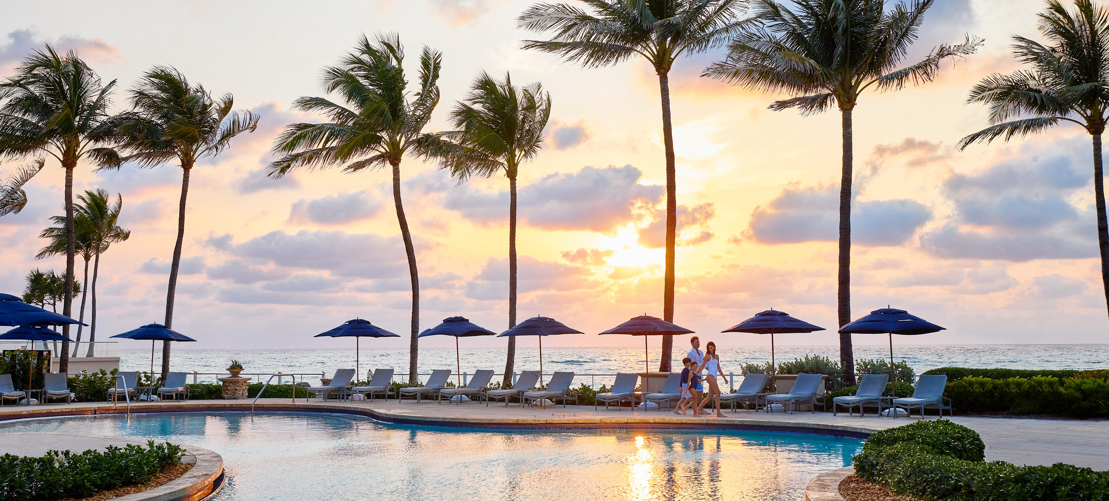 Tropical resort pool at sunset with palm trees, lounge chairs, umbrellas, and ocean view.