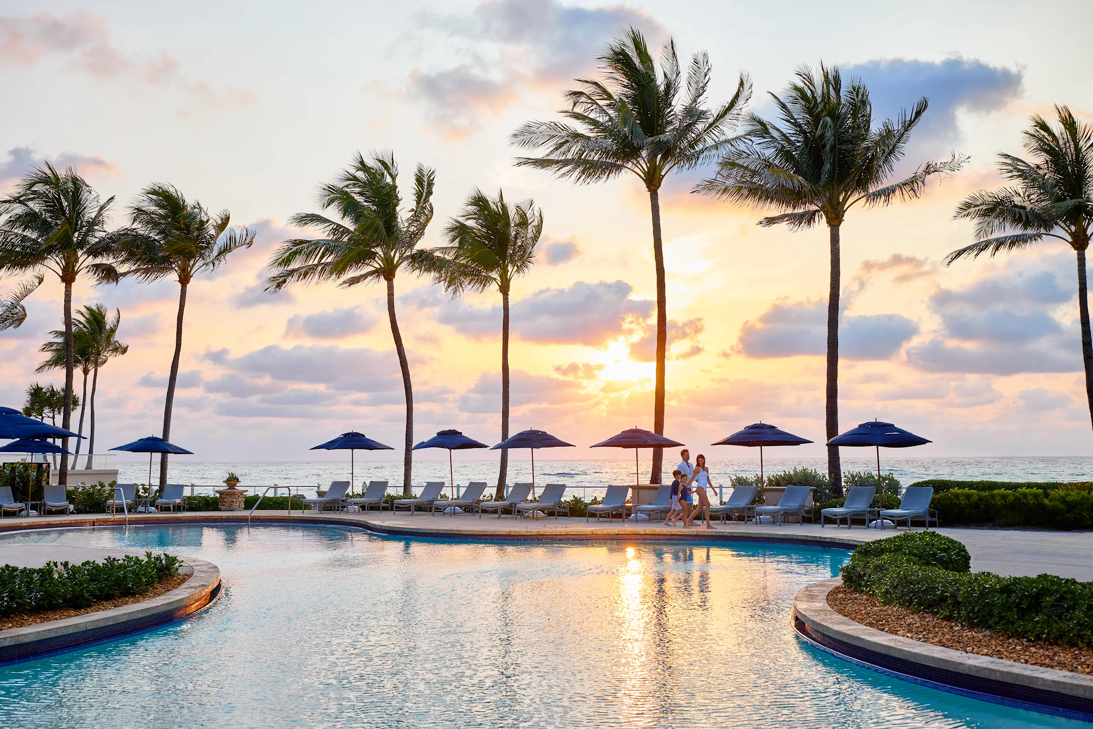 Tropical resort pool at sunset with palm trees, lounge chairs, umbrellas, and ocean view.