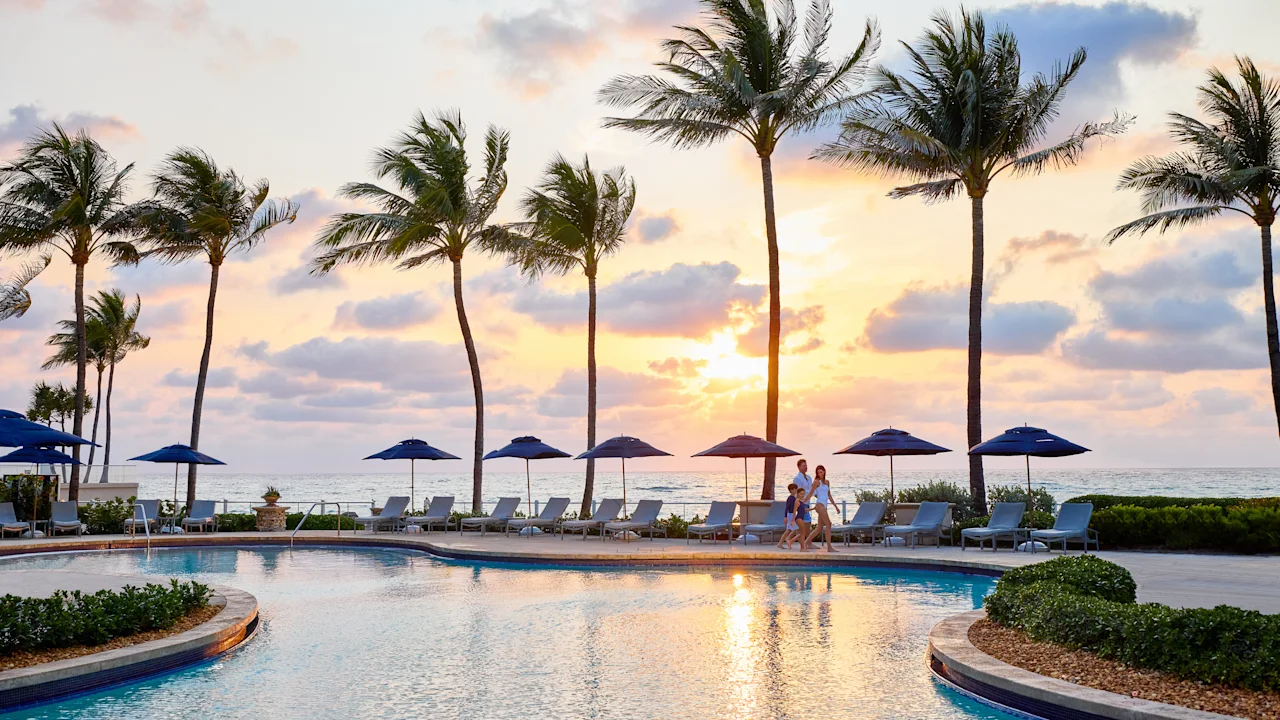 Tropical resort pool at sunset with palm trees, lounge chairs, umbrellas, and ocean view.
