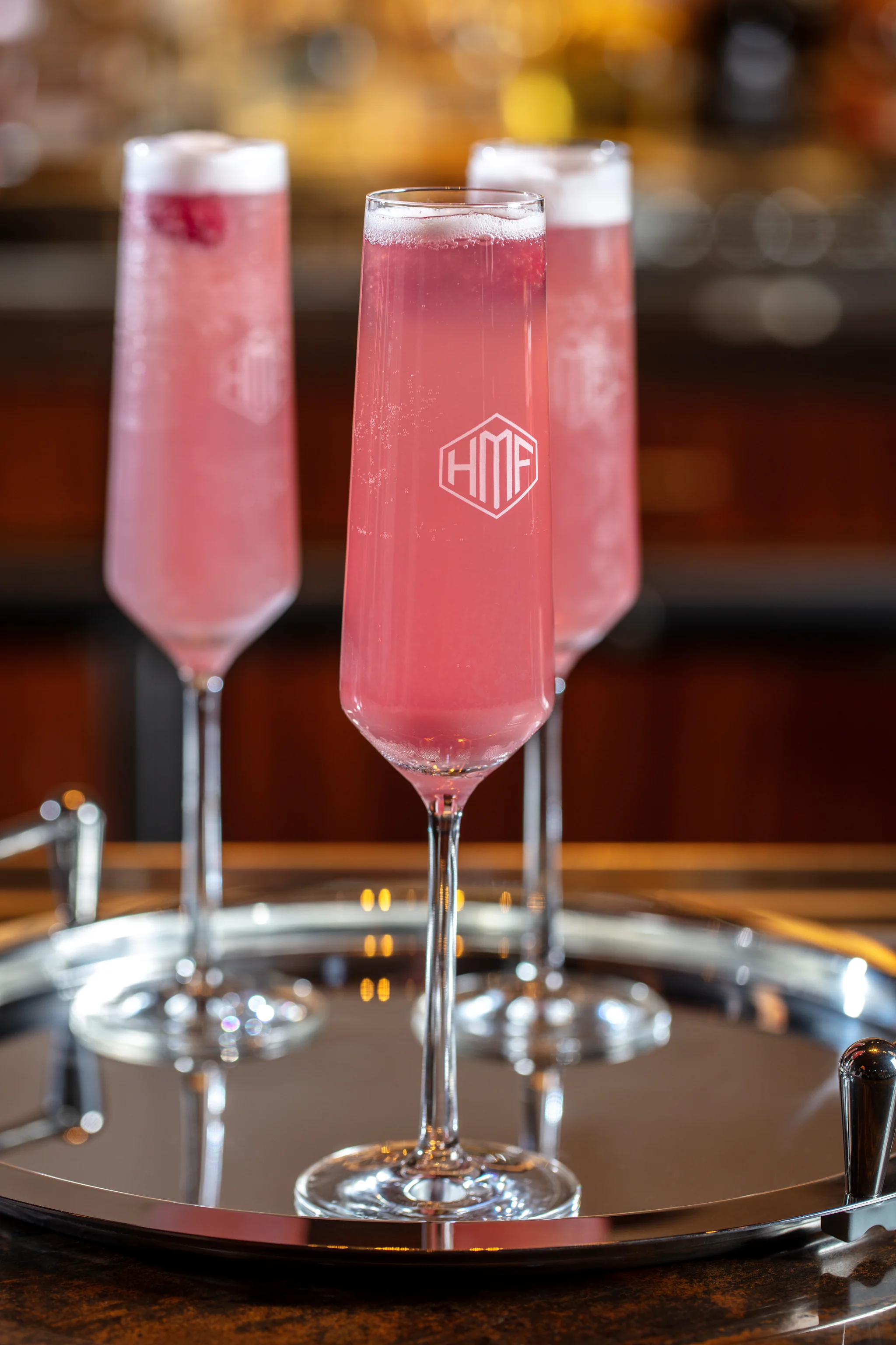 Three pink champagne cocktails with white foam tops in tall flute glasses on a silver tray.