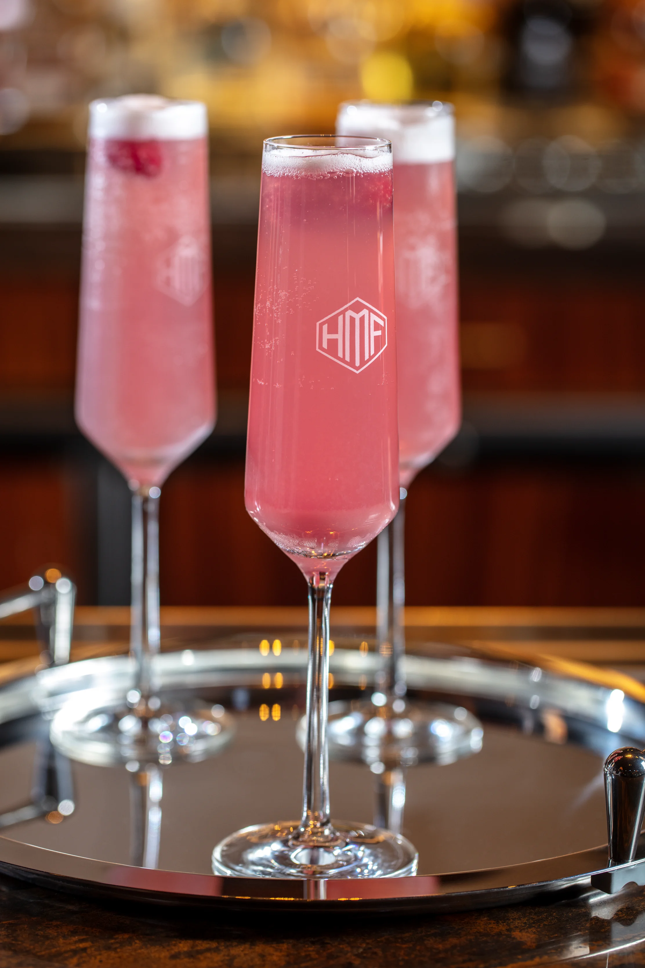 Three pink champagne cocktails with white foam tops in tall flute glasses on a silver tray.