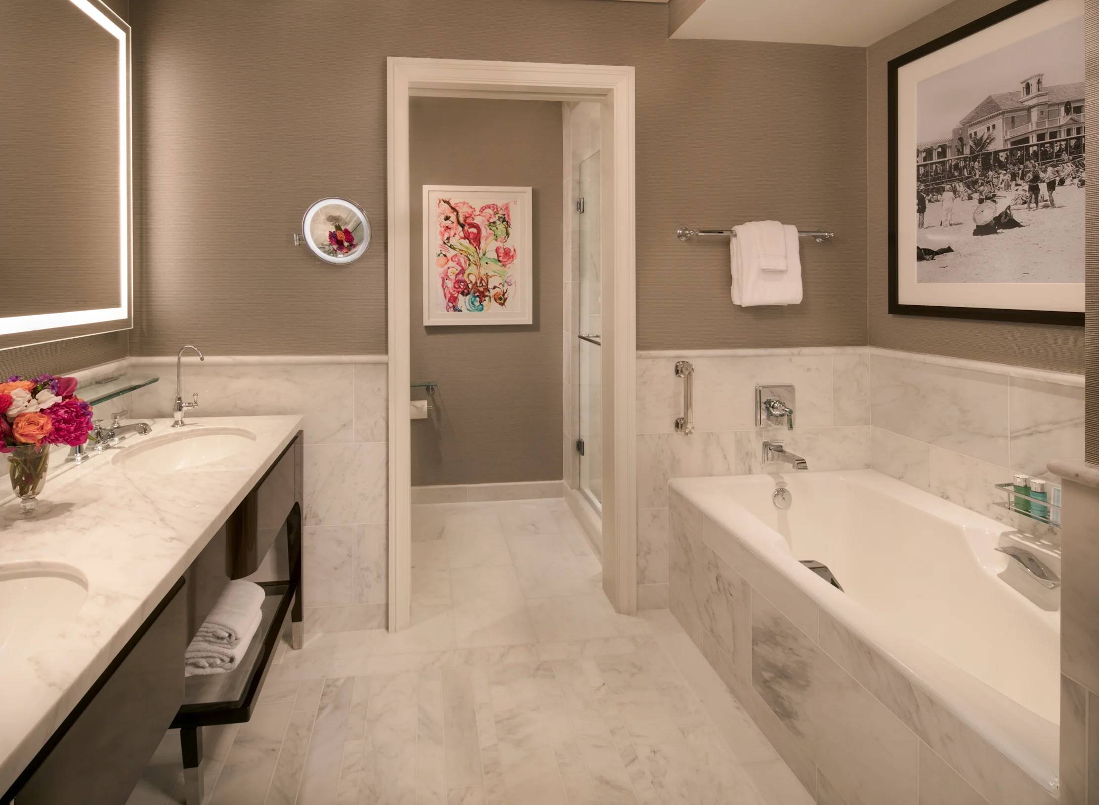 Modern hotel bathroom with white marble tub, double vanity, taupe walls, and floral artwork.