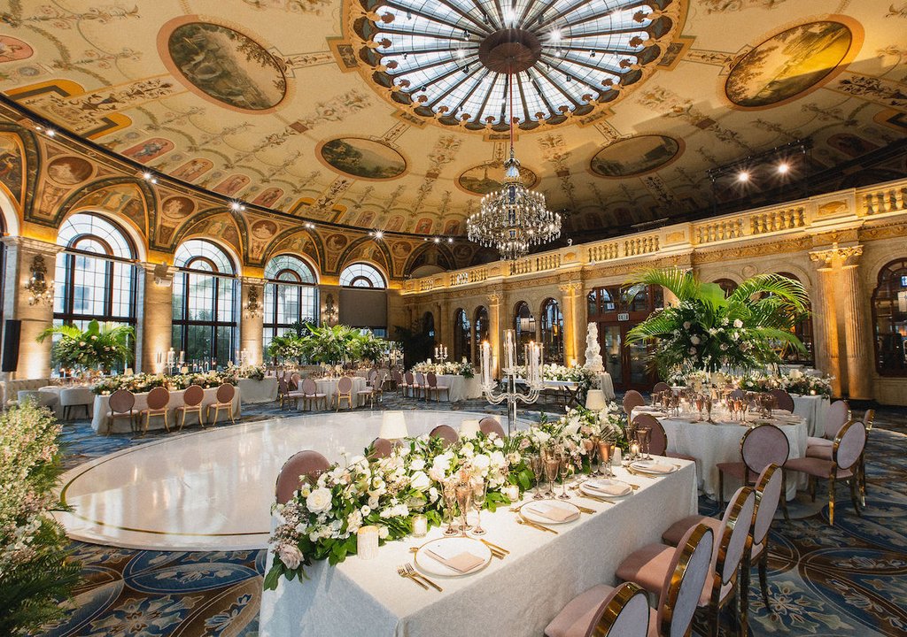 Elegant ballroom with ornate painted ceiling, crystal chandelier, and formal dining tables with floral centerpieces.